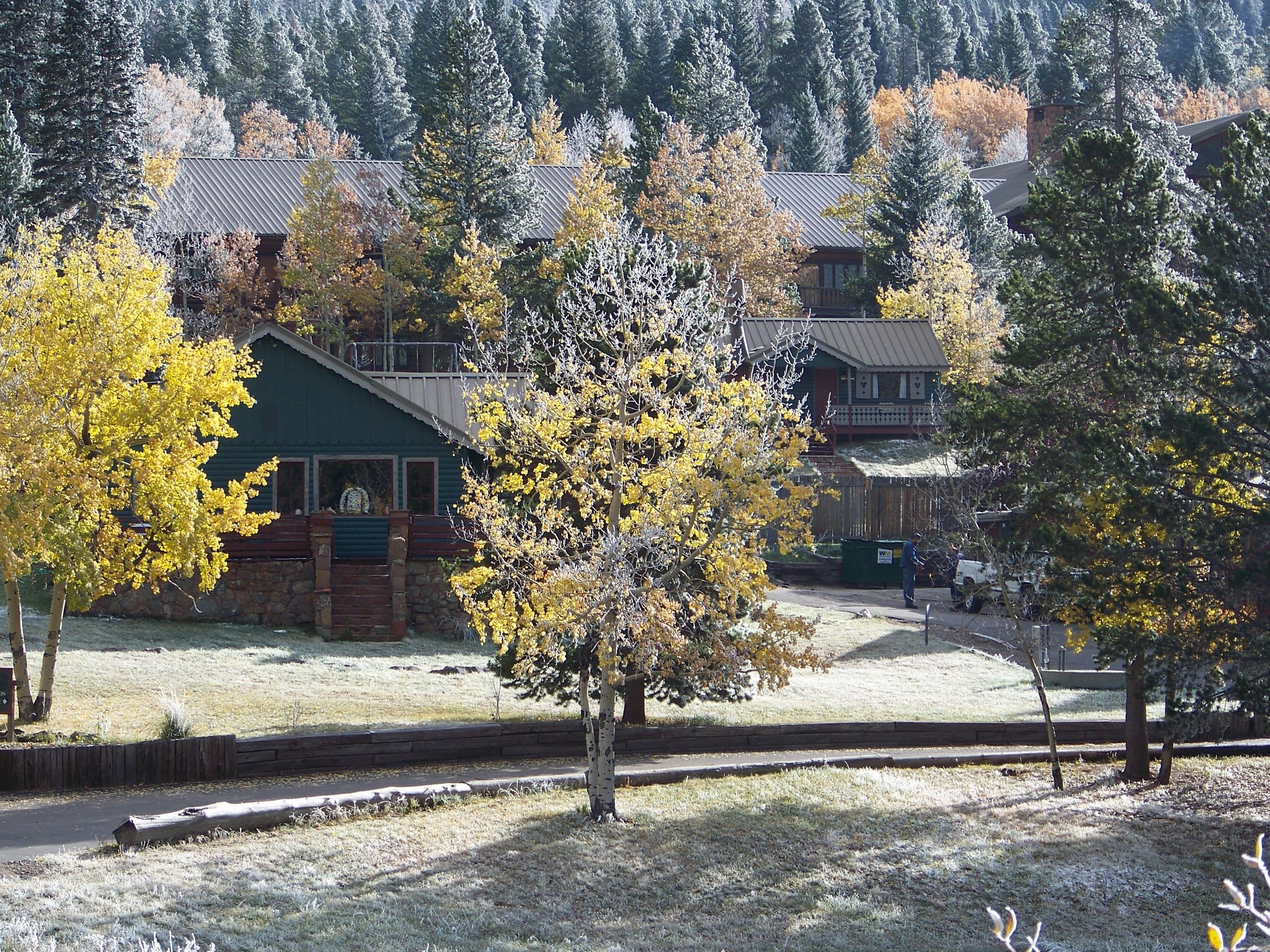 A rustic lodge with a stone exterior and a green roof, surrounded by autumn-colored trees and a mountainous backdrop.