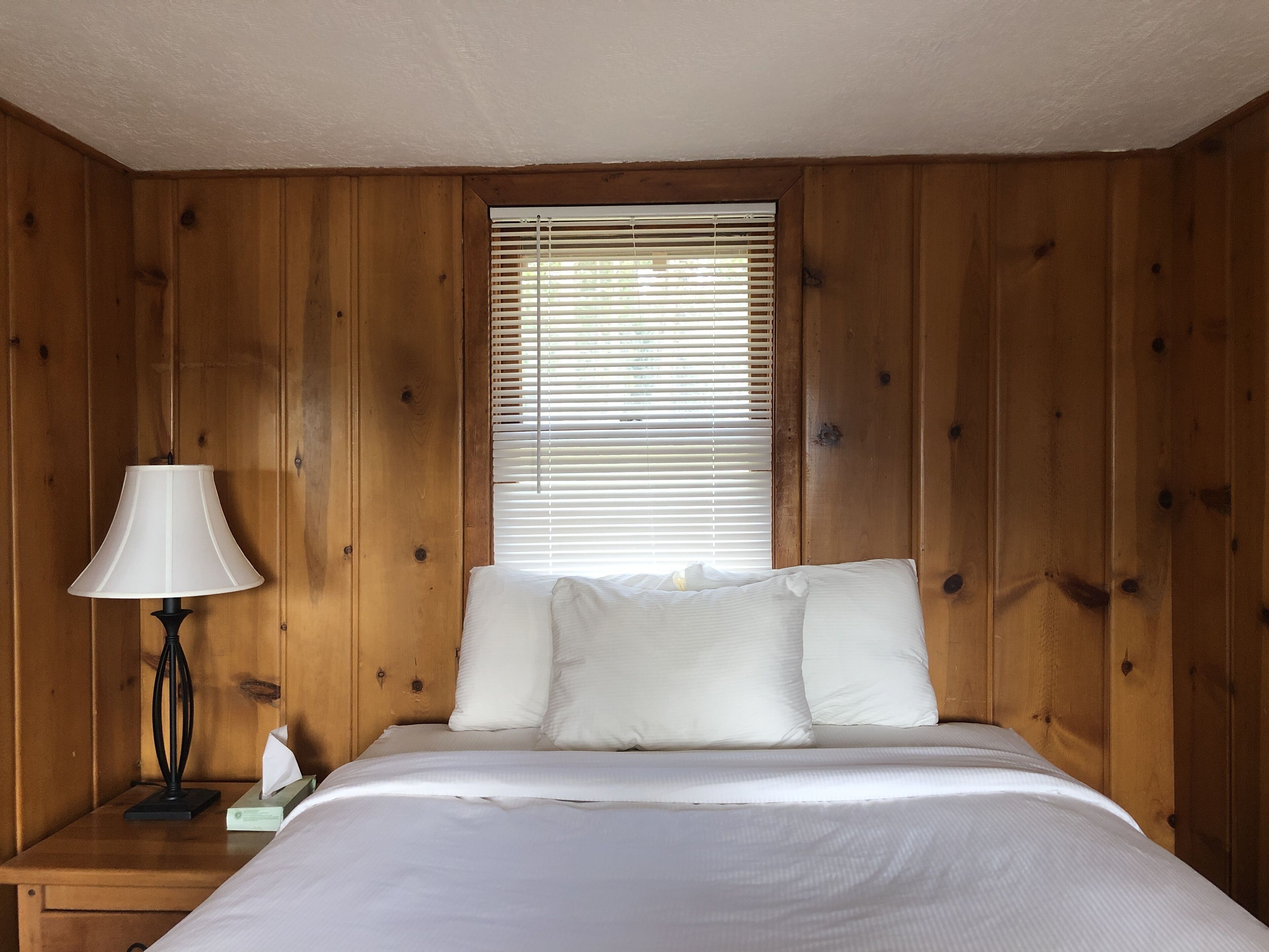A room with wooden panel walls, a bed with white bedding, a bedside lamp, and a window with blinds.