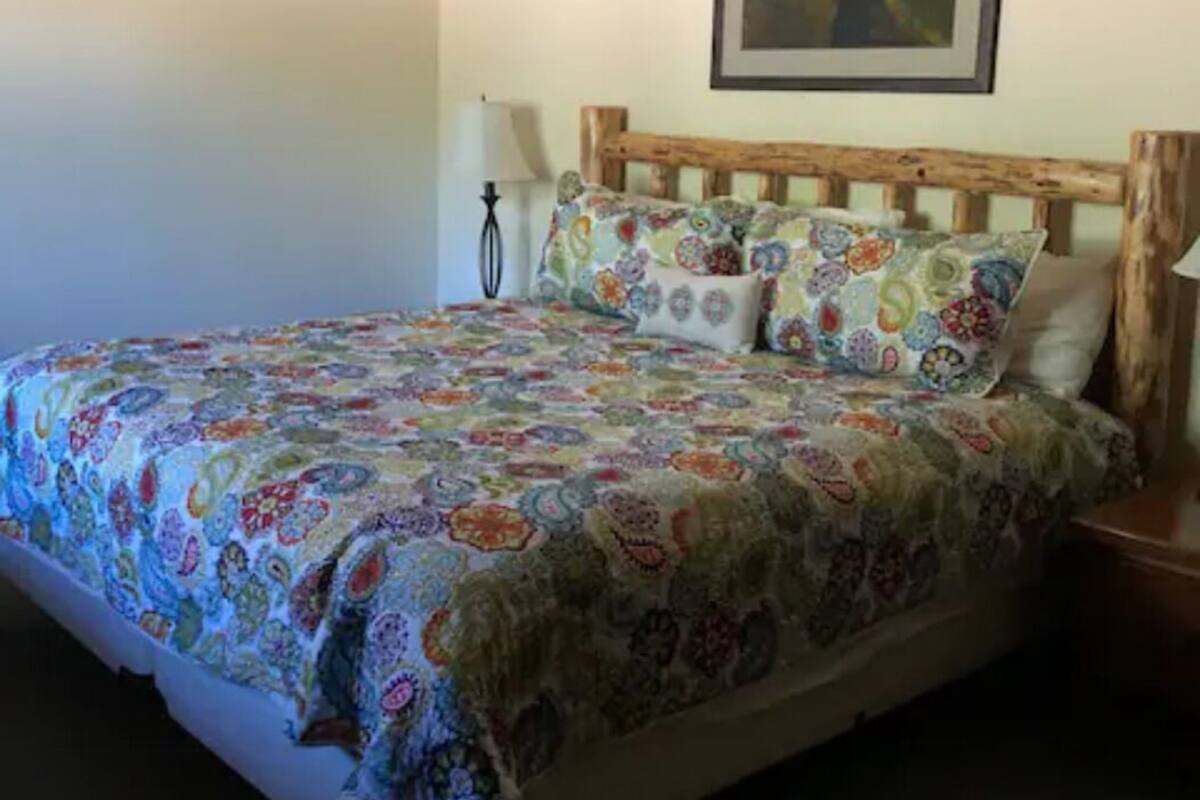 A bed with a colorful quilt, a wooden headboard, a framed picture on the wall, and a bedside lamp.