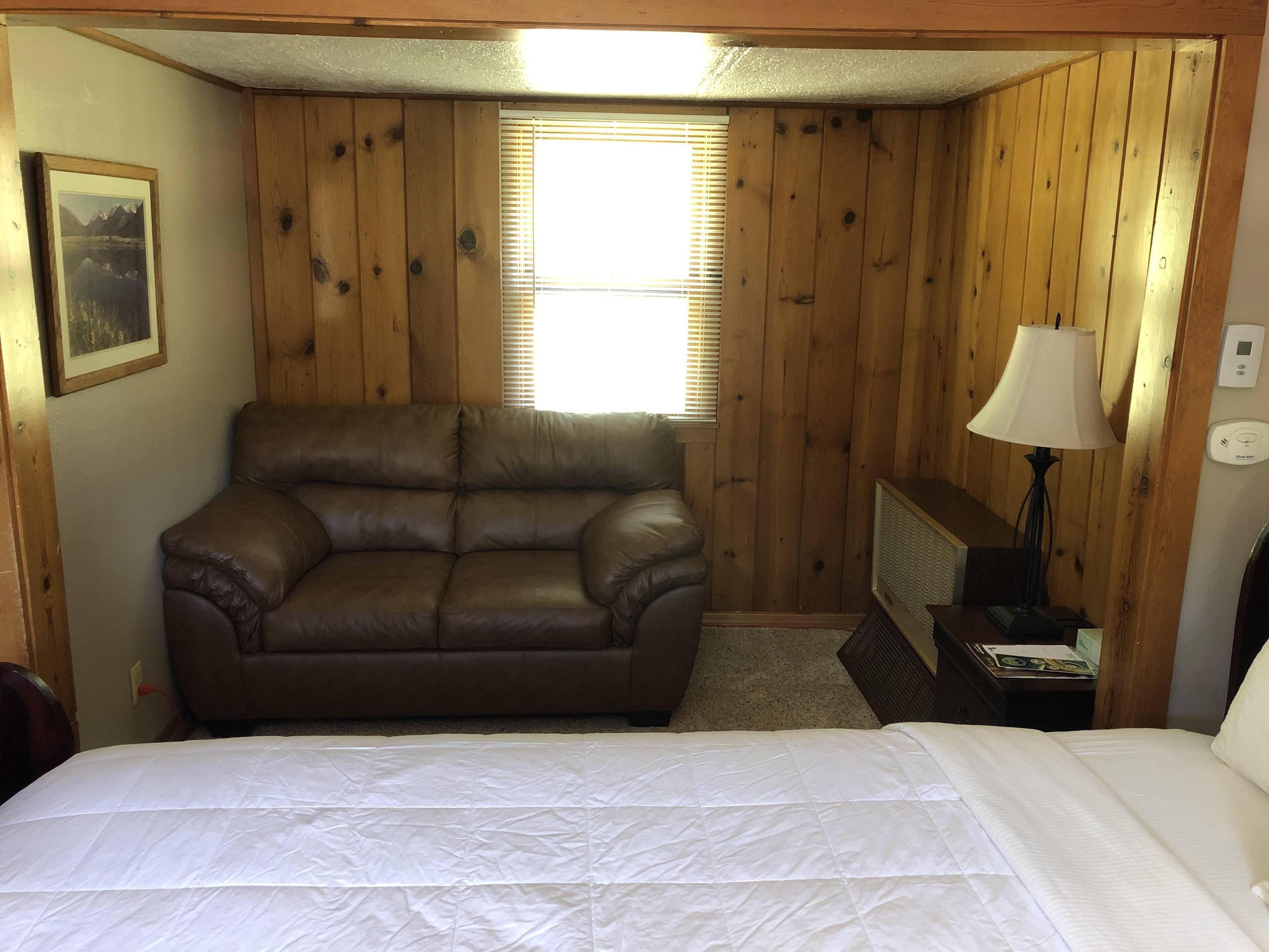 A room with a brown leather sofa, a white bed, wooden walls, a window with blinds, a lamp, and a thermostat.