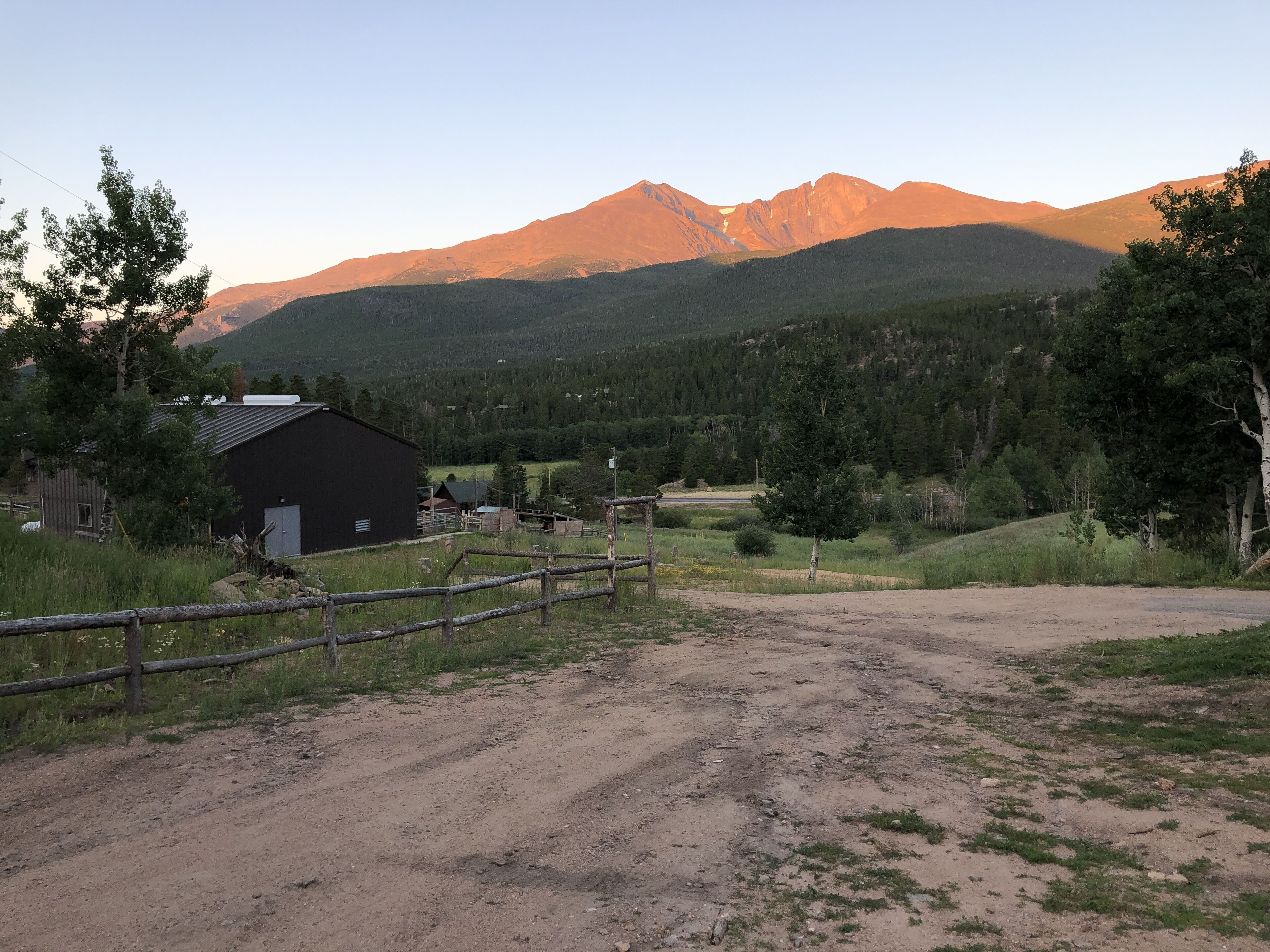 A rural landscape with a dirt road leading to a black building, surrounded by trees and mountains in the background.