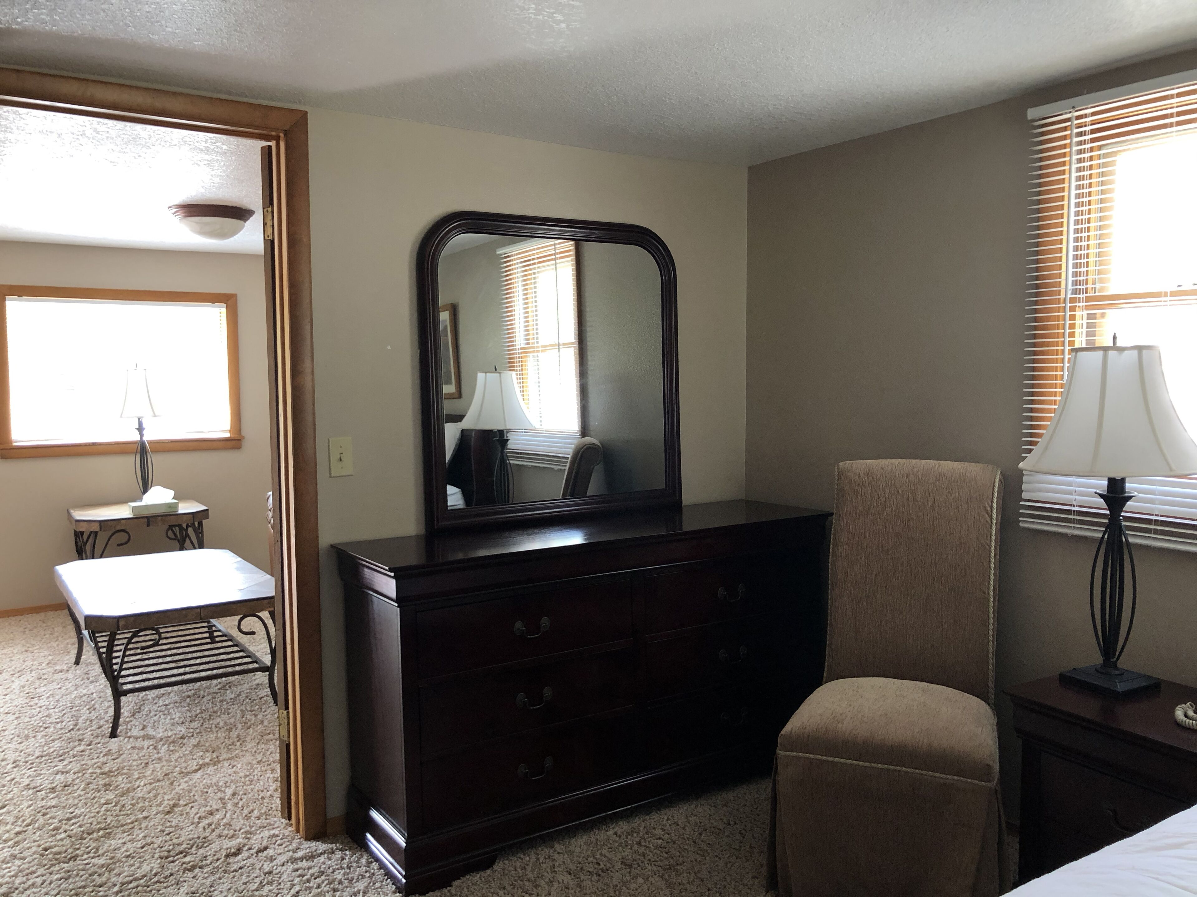 A room with a wooden dresser, a chair, a desk with a lamp, and a window with blinds.