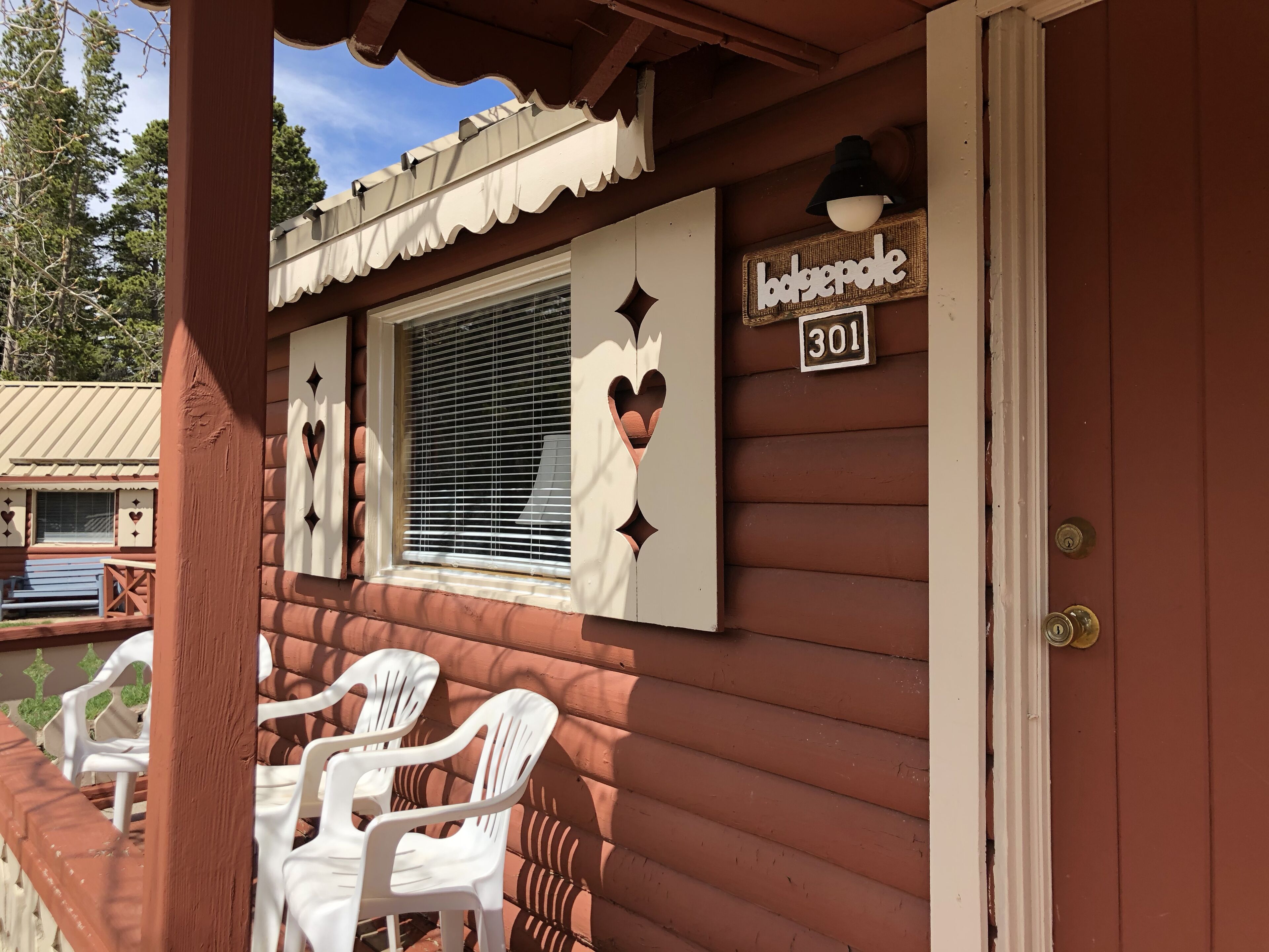 A wooden cabin with a balcony, white plastic chairs, a window with blinds, and a sign reading "bodgepole 301".