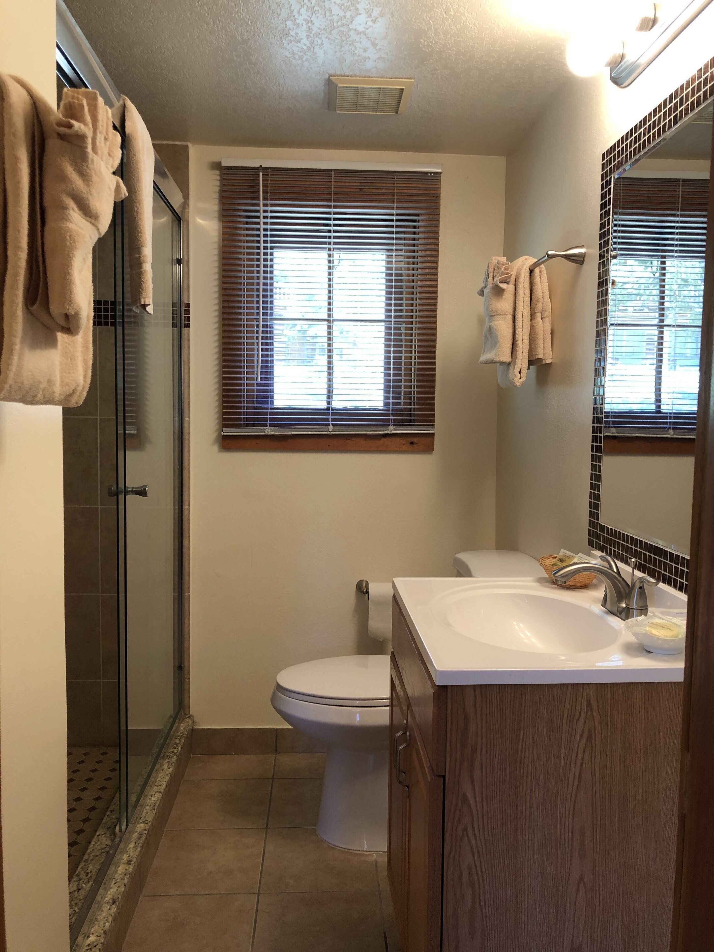A bathroom with a glass shower enclosure, a toilet, a sink with a mirror, and a window with blinds.