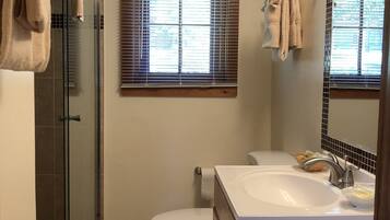 A bathroom with a glass shower enclosure, a toilet, a sink with a mirror, and a window with blinds.