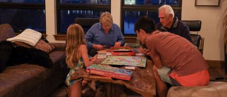 A group of people playing board games around a wooden table in a cozy room with large windows.