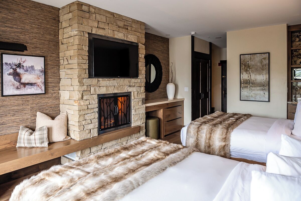 Une chambre d’hôtel avec une cheminée en pierre, un téléviseur fixé au mur, un lit recouvert d’un plaid en fourrure et un tableau représentant un élan.