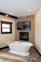 Un bagno con una vasca da bagno indipendente, un muro in pietra, una televisione montata sopra un camino e una finestra con persiane.