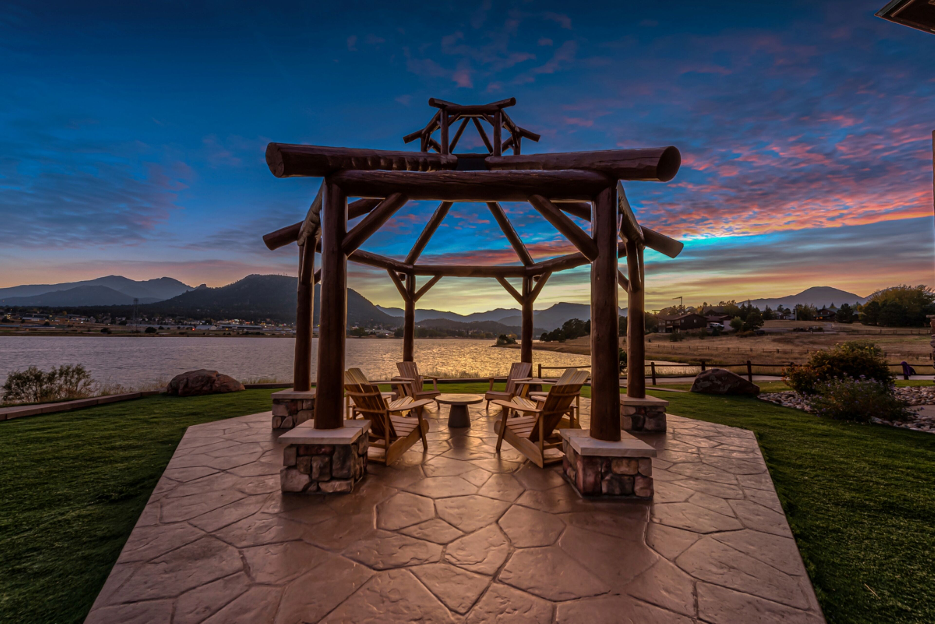 A gazebo with a stone floor and wooden chairs, overlooking a lake with mountains in the background.