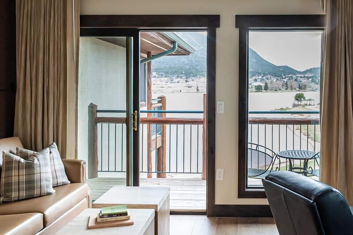 A room with a sofa, a small table, a chair, and a balcony with a view of a beach and mountains.