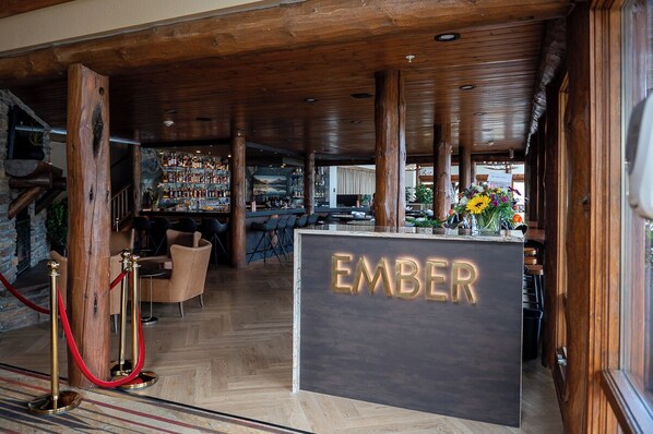 A bar area with a wooden ceiling, a reception desk labeled 'EMBER', and a bar stocked with bottles.