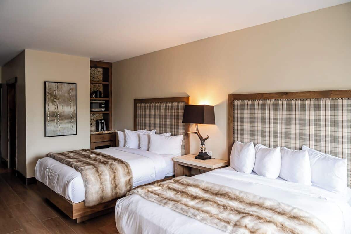 A hotel room with two beds, a headboard with a plaid pattern, a bedside table with a lamp, and a wall-mounted shelf with books and decorative items.
