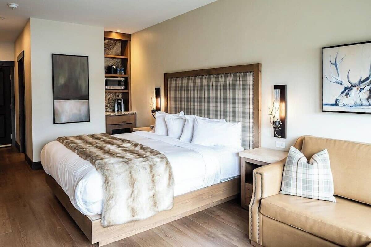 A modern hotel room with a large bed, a wooden nightstand, a leather sofa, and a wall-mounted shelf with books and decorative items.