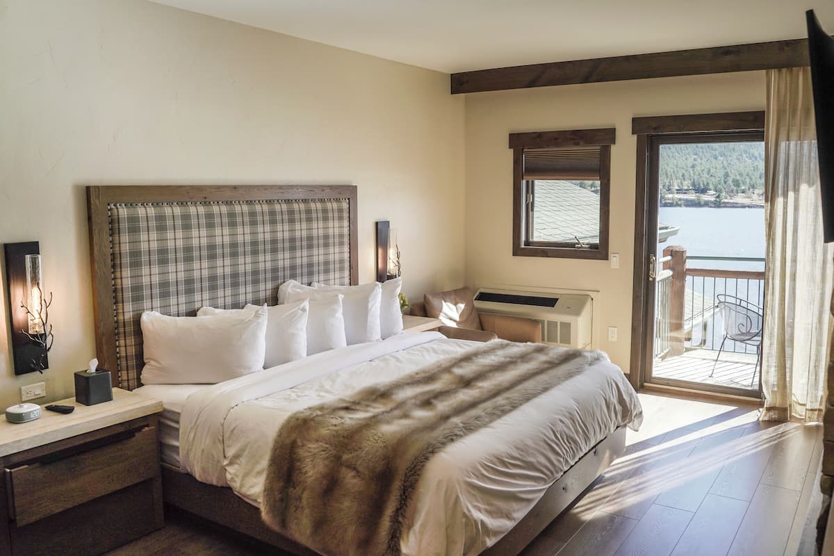 A bedroom with a large bed, a bedside table with a lamp, a window with a view of a body of water, and a balcony with a chair.