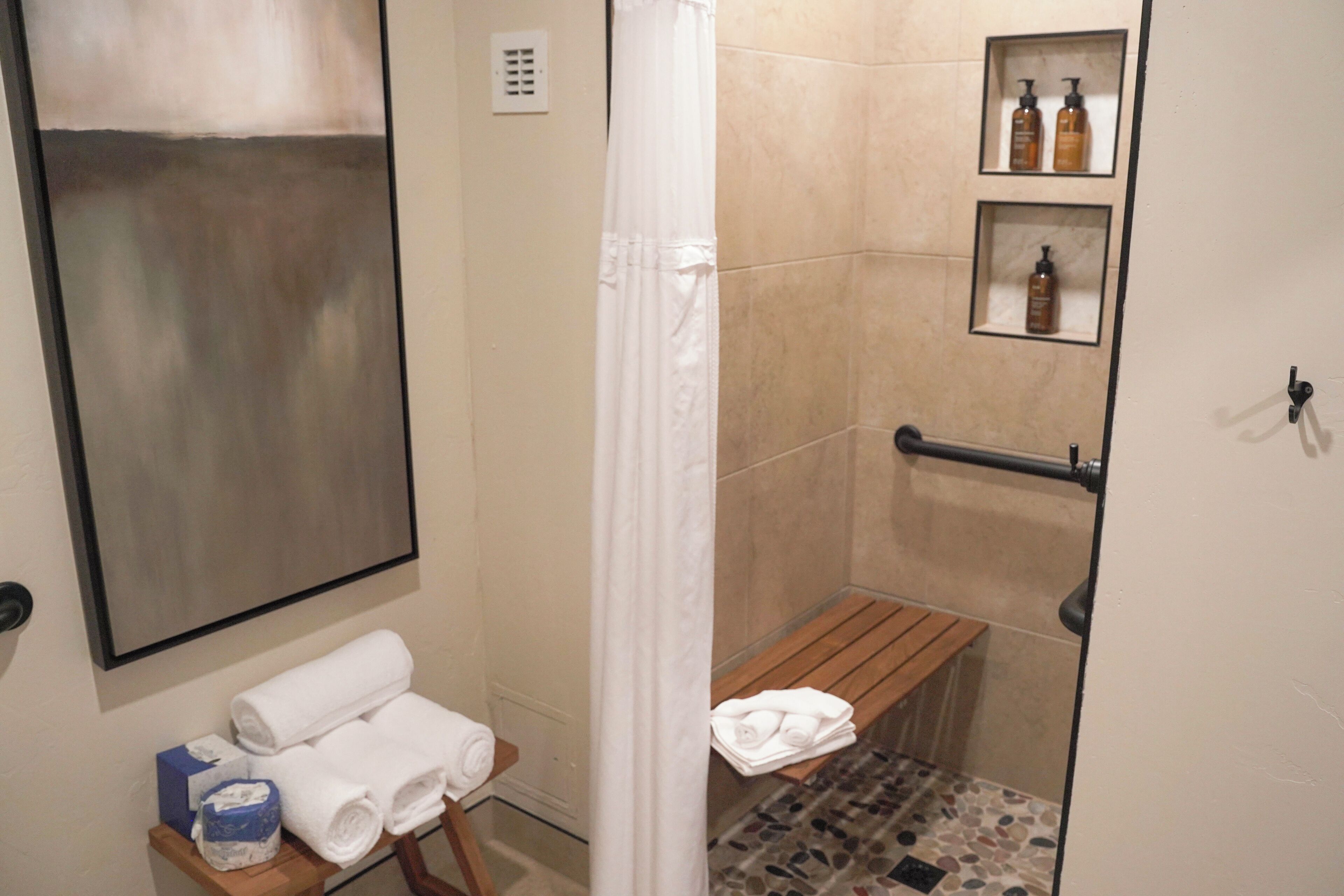 A bathroom with a shower, a bench, a towel rack, and a shelf with toiletries.