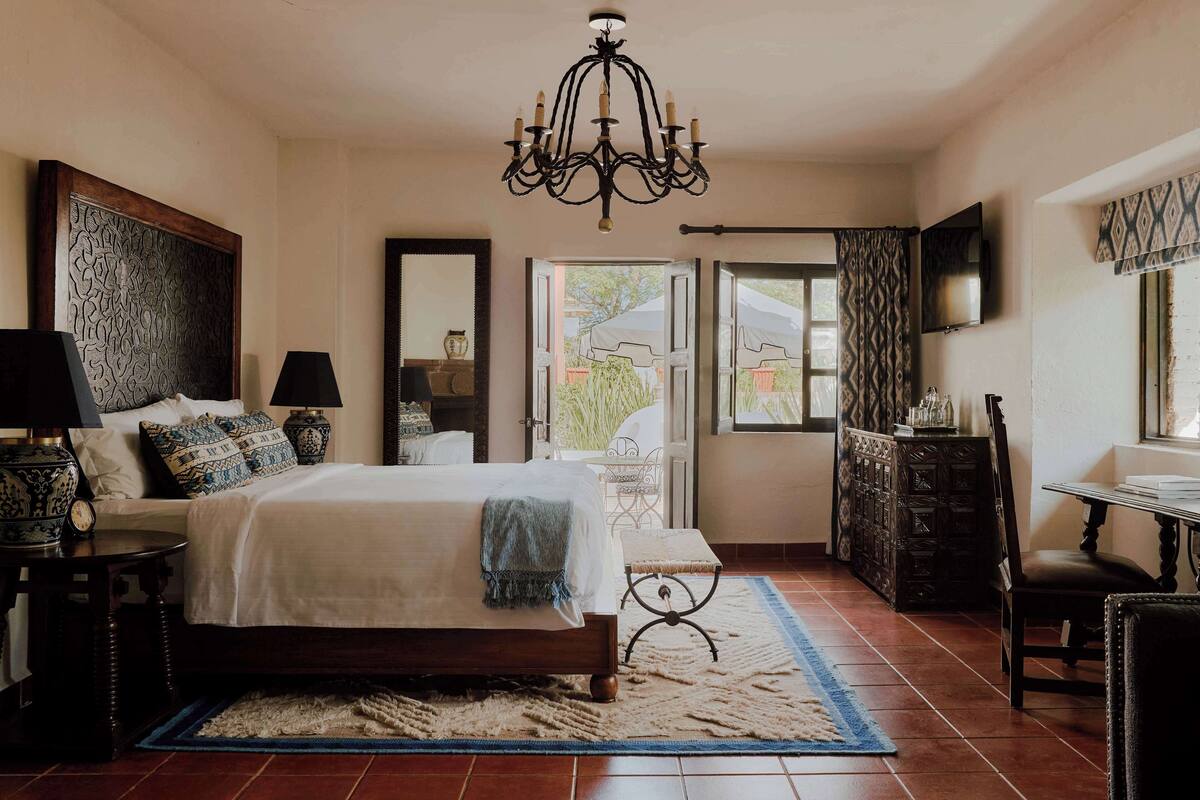 A bedroom with a large bed, a chandelier, a wooden headboard, a bedside table with lamps, a mirror, a window with curtains, and a view of the outside.