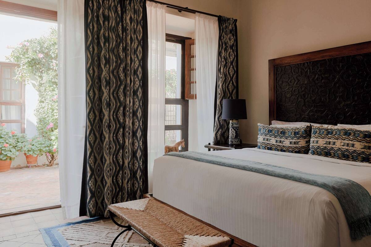 A bedroom with a bed, a bench, a nightstand with a lamp, patterned curtains, and a view of the outside.