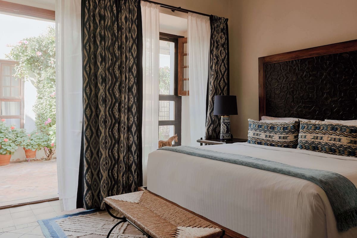 Un dormitorio con cama, un banco, una mesita de noche con lámpara, cortinas estampadas y vistas al exterior.