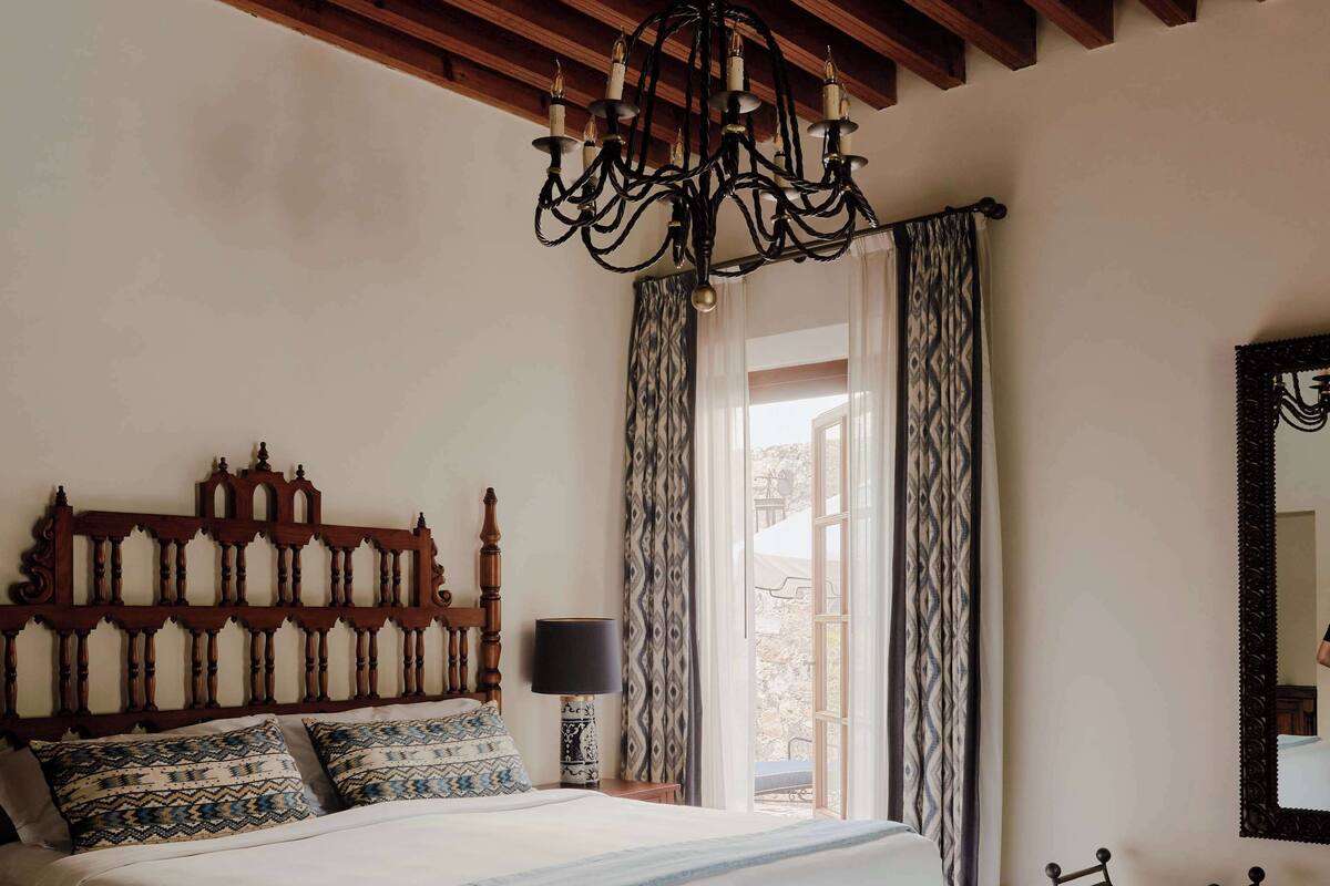 A bedroom with a large bed, a wooden headboard, a chandelier, a nightstand, a chair, and a window with curtains.