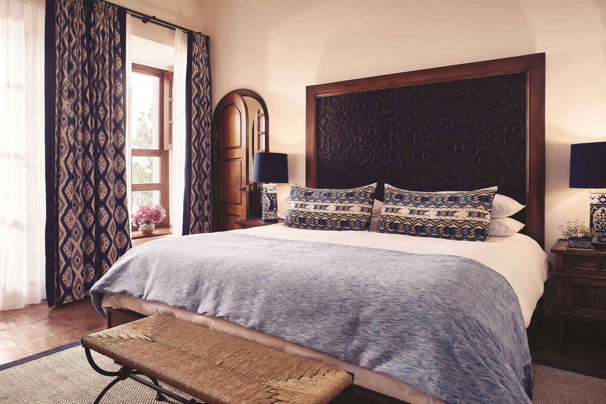 A bedroom with a large bed, patterned pillows, a wooden headboard, a bedside table with a lamp, and a bench.