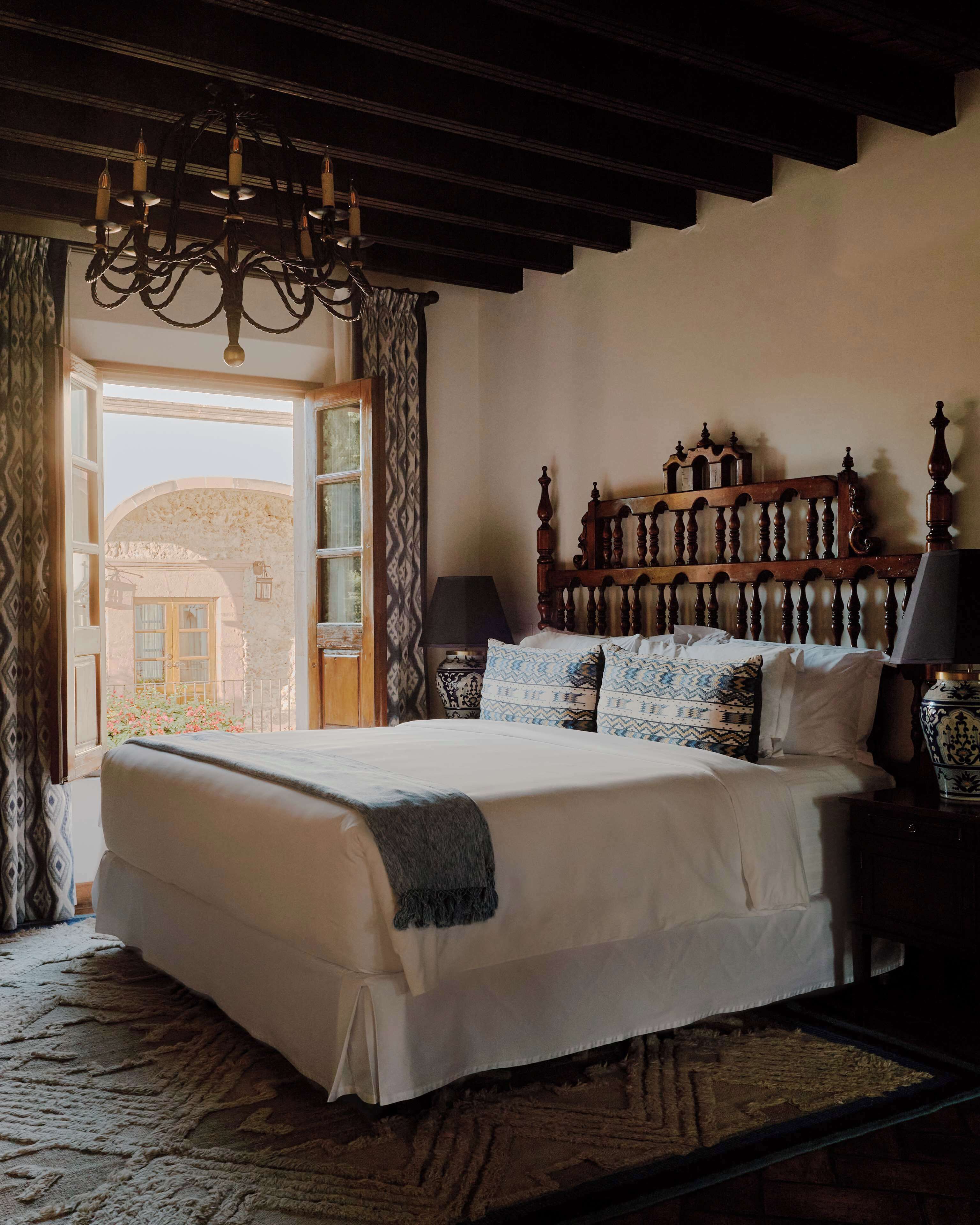 A bedroom with a large bed, a wooden headboard, a chandelier, a nightstand, and a view of the outside.