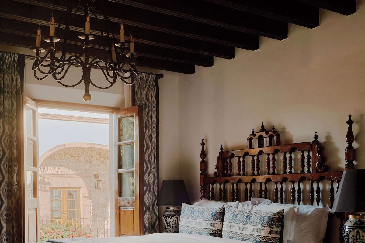 A bedroom with a large bed, a wooden headboard, a chandelier, a nightstand, and a view of the outside.