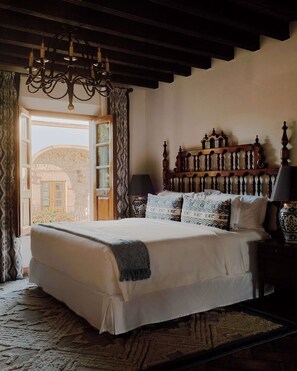 A bedroom with a large bed, a chandelier, a wooden headboard, a nightstand, and a view of the outside.