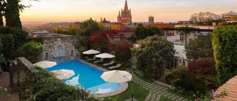 A pool area with umbrellas, surrounded by greenery and a view of a historic building in the distance.
