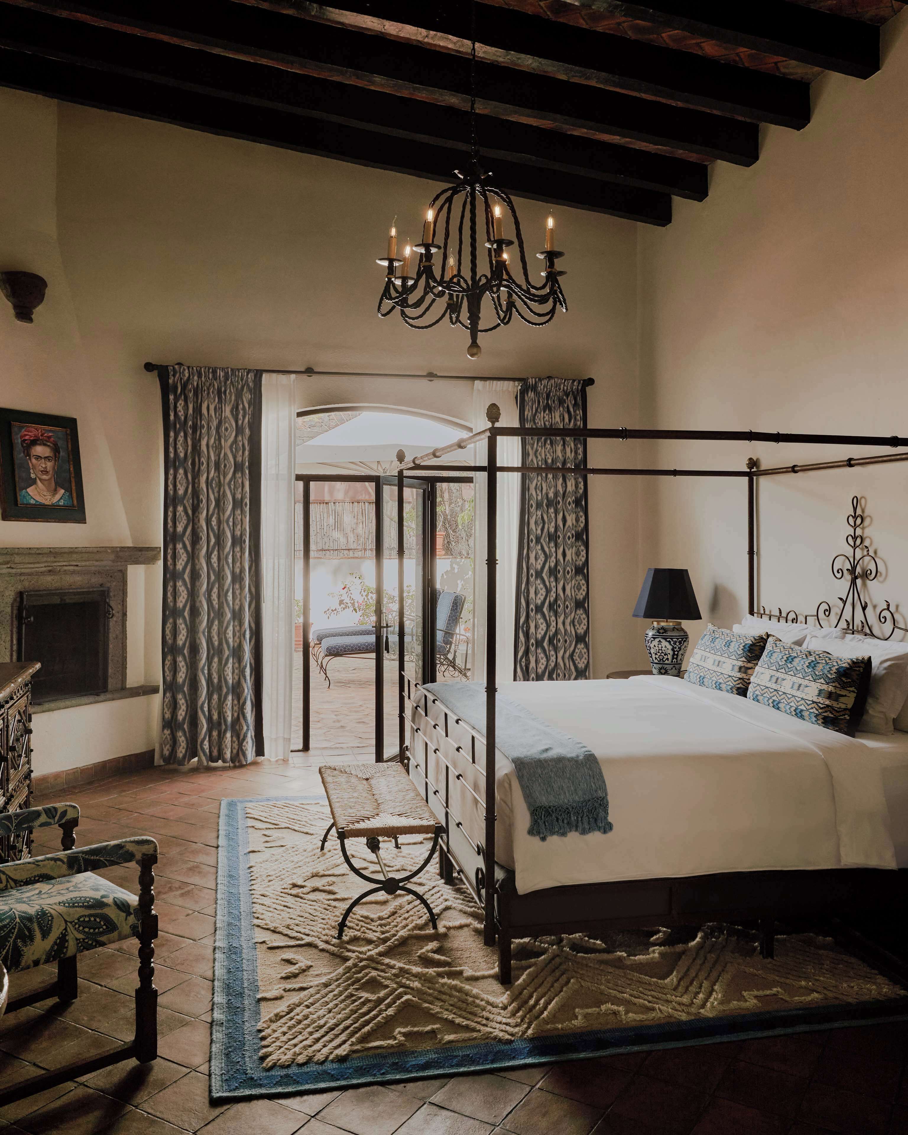A bedroom with a four-poster bed, a chandelier, a fireplace, and a view through a glass door to an outdoor patio.