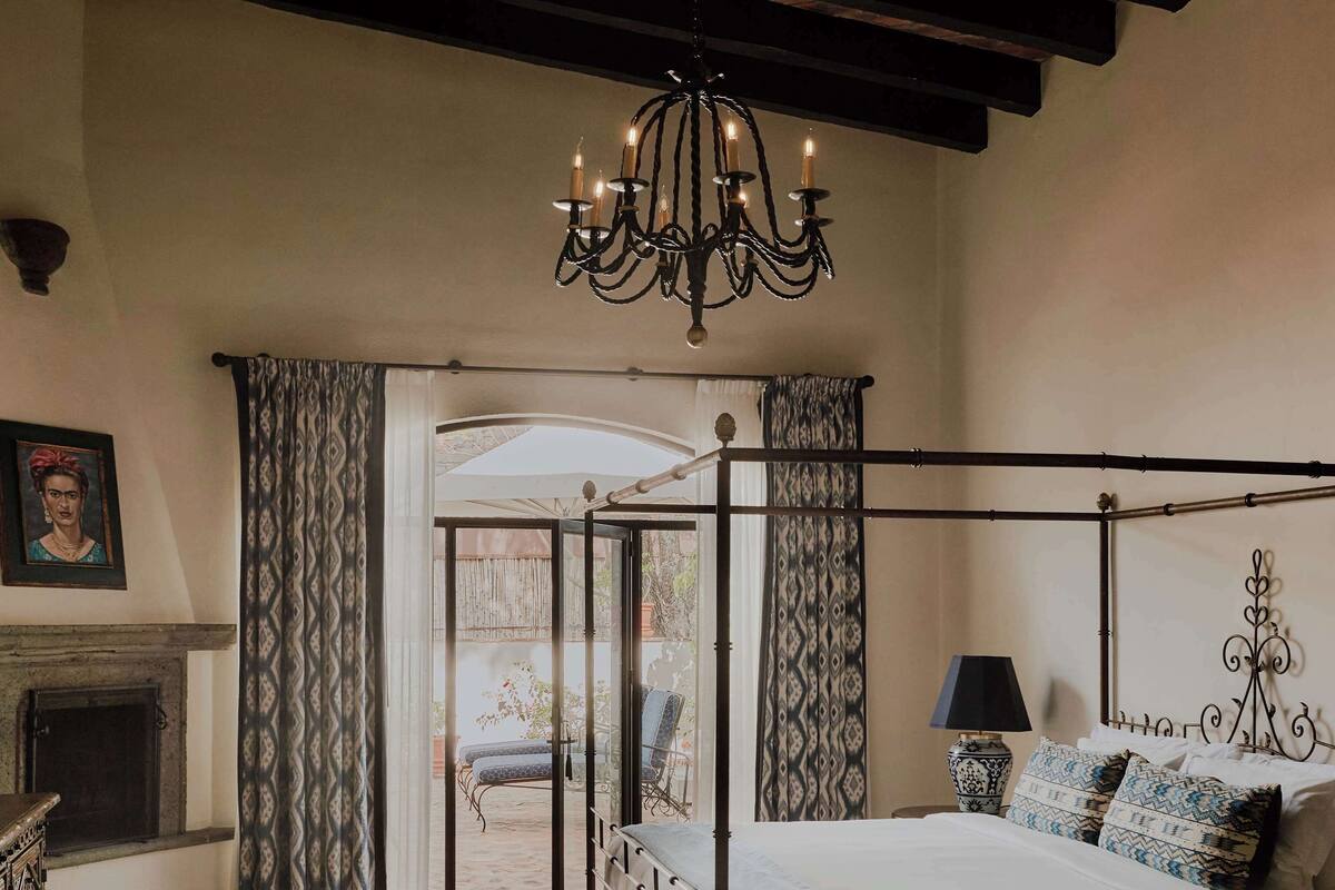 A bedroom with a four-poster bed, a chandelier, a fireplace, and a view through a glass door to an outdoor patio.
