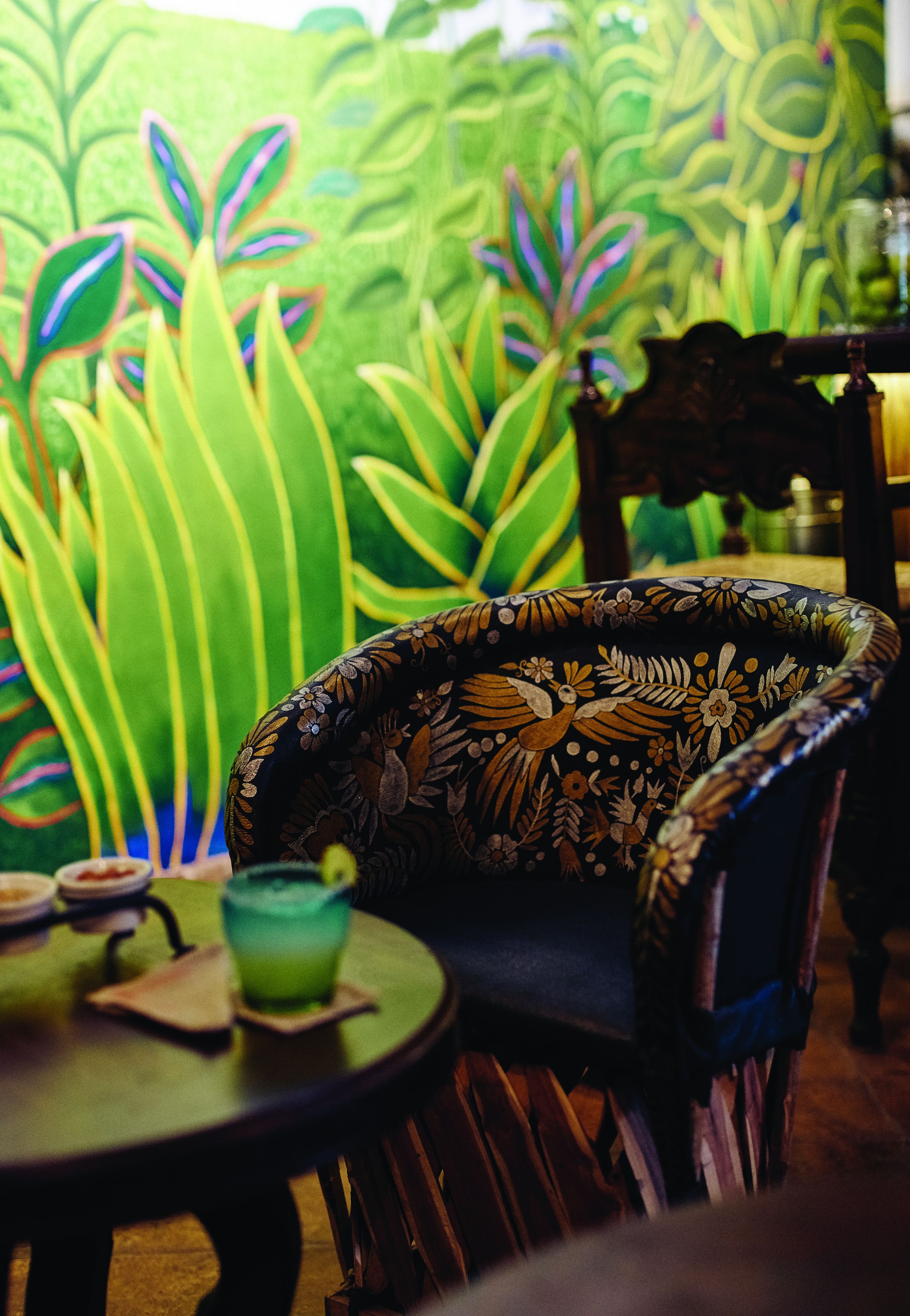 Una silla con estampado floral, una mesa con un vaso de vidrio verde, y un mural con follaje verde de fondo.