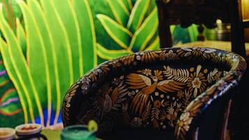 Una silla con estampado floral, una mesa con un vaso de vidrio verde, y un mural con follaje verde de fondo.