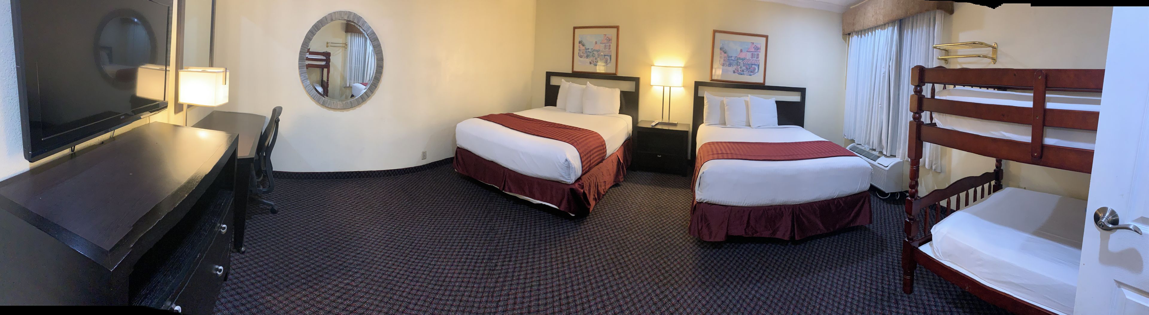 A hotel room with two single beds, a desk with a chair, a large mirror, and a bathroom with a bathtub and shower.