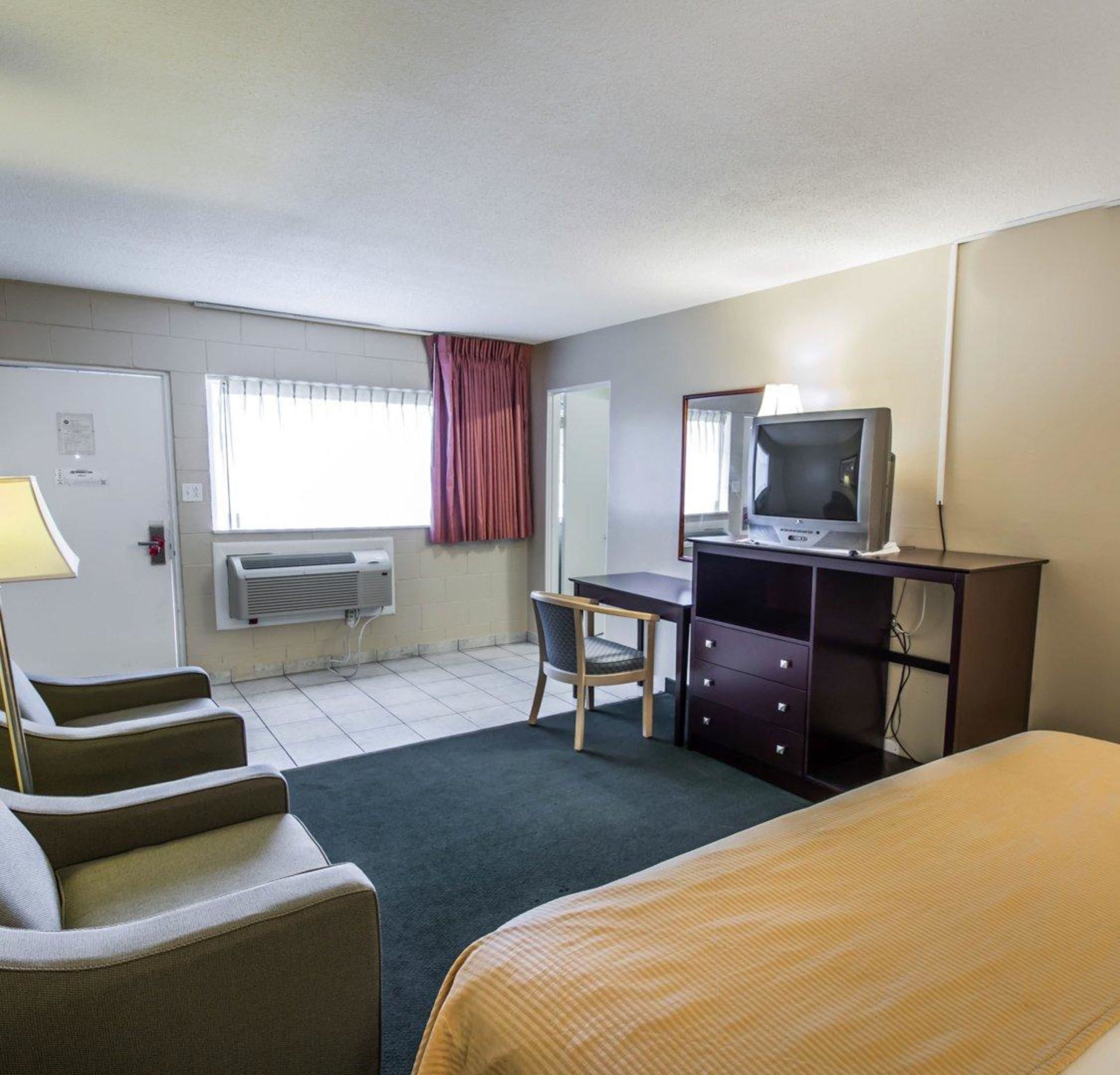 A hotel room with a bed, a desk with a computer, a chair, a TV, and an air conditioning unit.