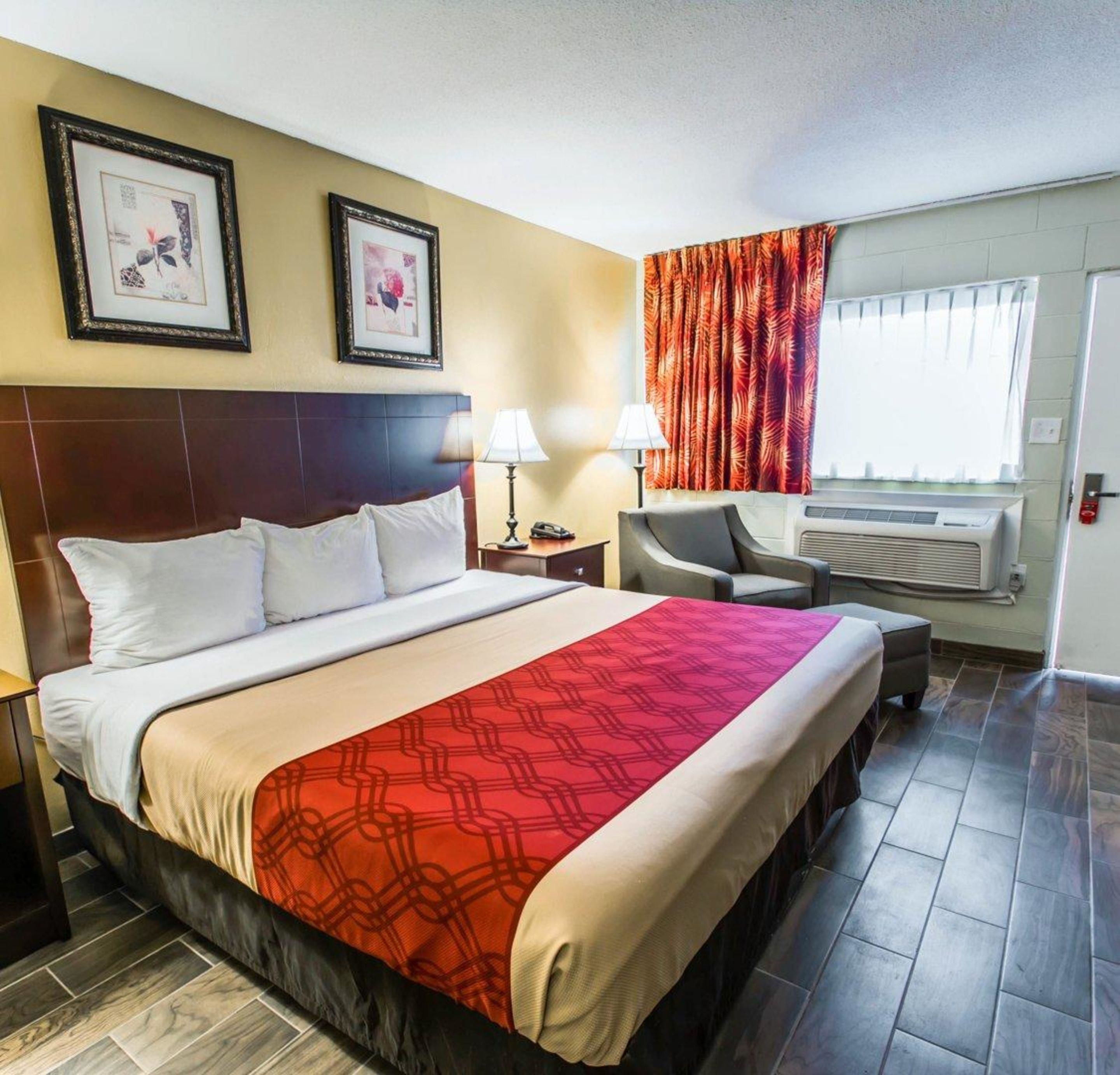 Ein Hotelzimmer mit einem großen Bett, zwei Nachttischen, einem Sessel, einem Schreibtisch, einem Fenster mit Vorhängen und einer Wand-Klimaanlage.