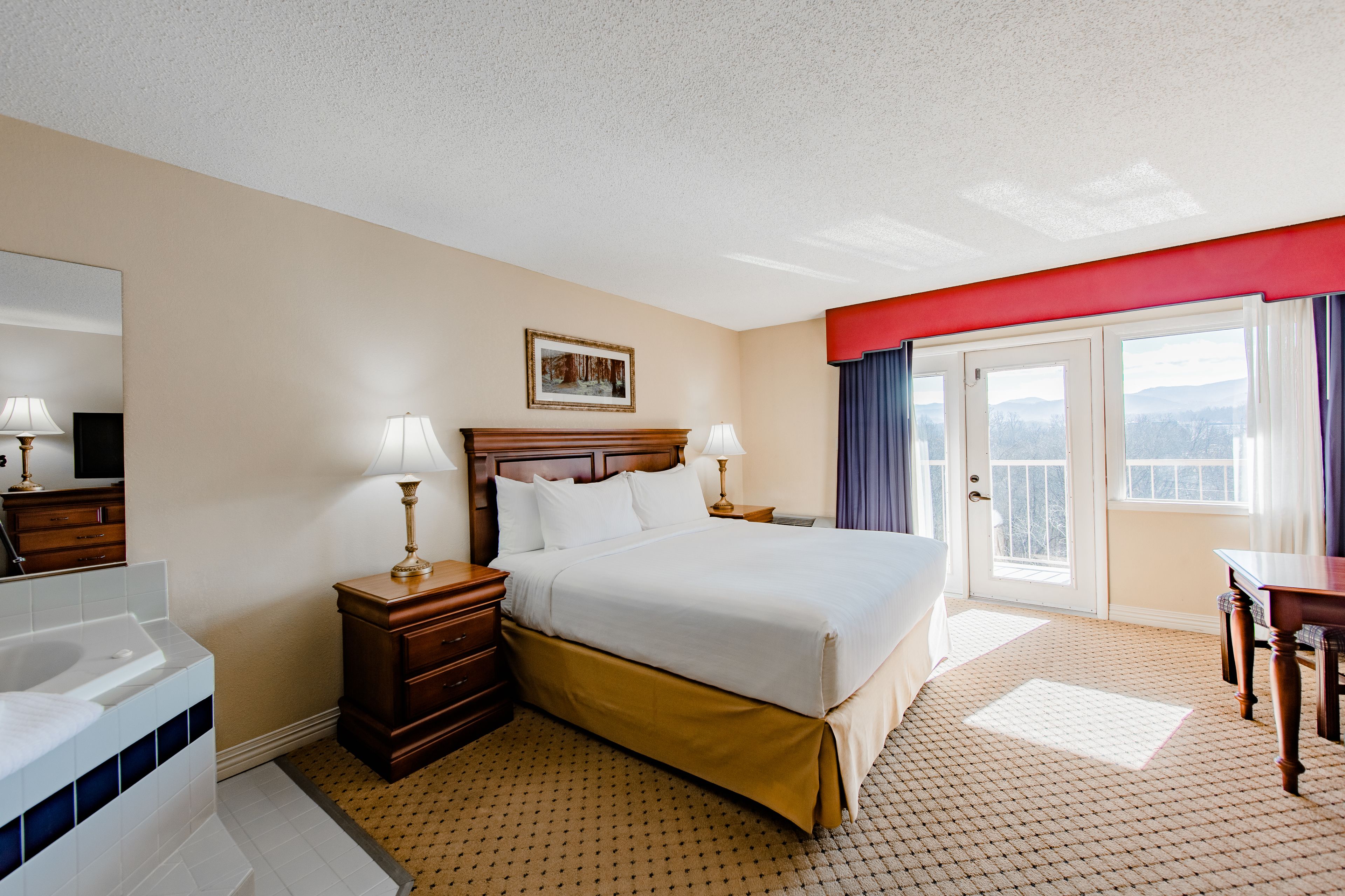 A hotel room with a large bed, a wooden nightstand, a bathroom with a bathtub, and a balcony with a view.