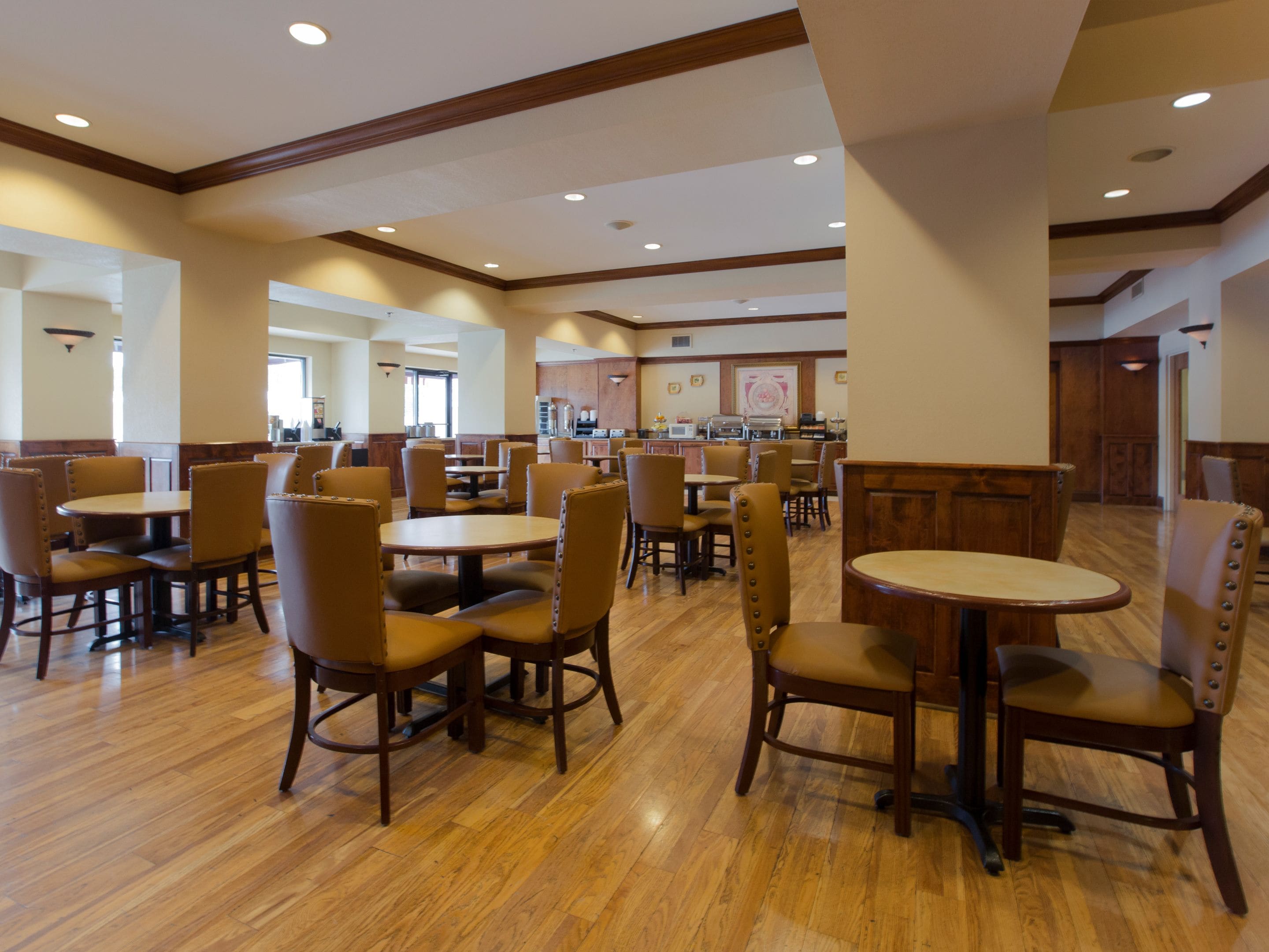 A spacious dining area with multiple tables and chairs, a kitchen in the background, and a wooden floor.