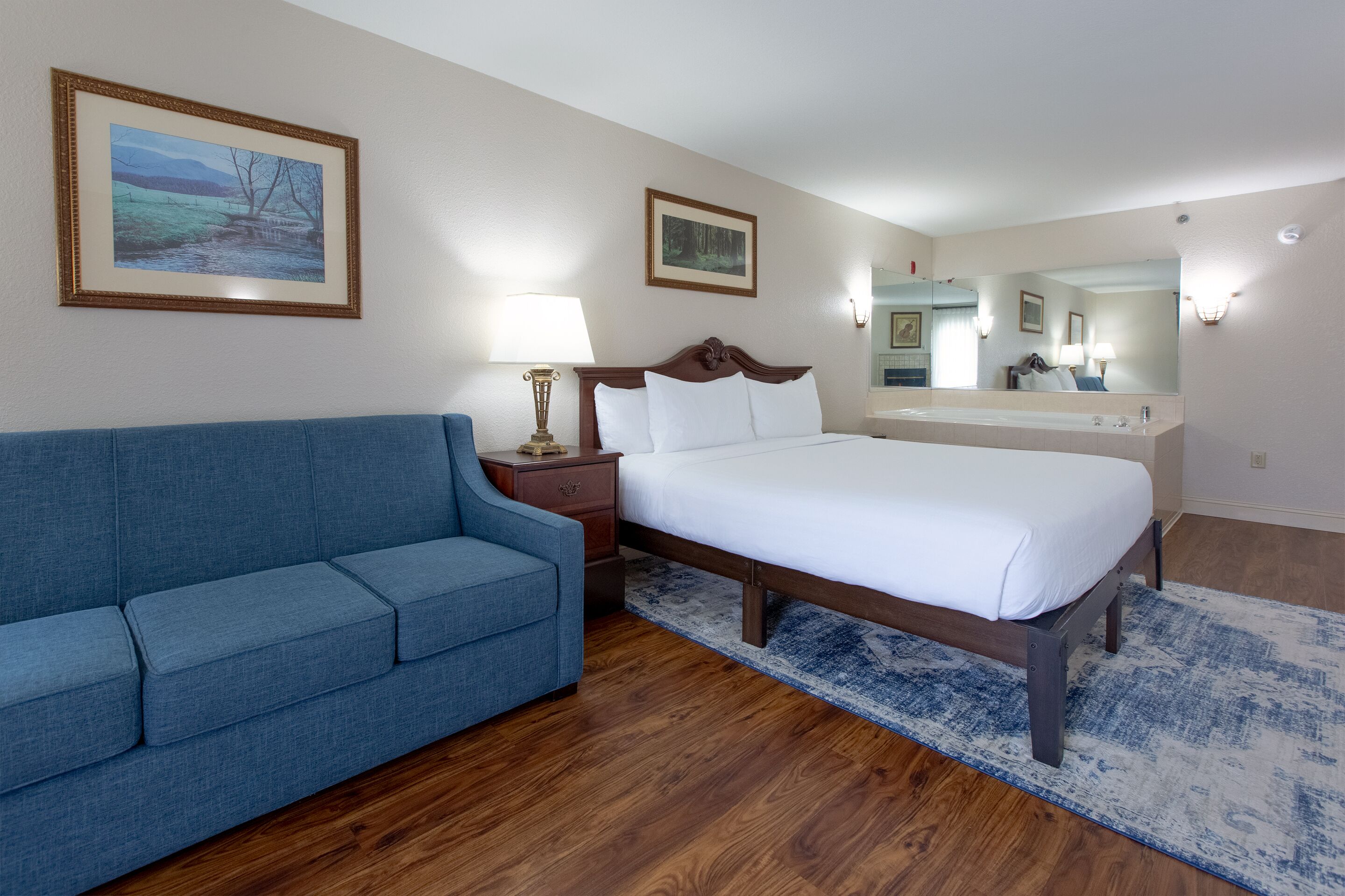 Hotelkamer met een bed, een blauwe bank, een houten vloer, een badkamer met een grote spiegel en twee ingelijste foto's aan de muur.