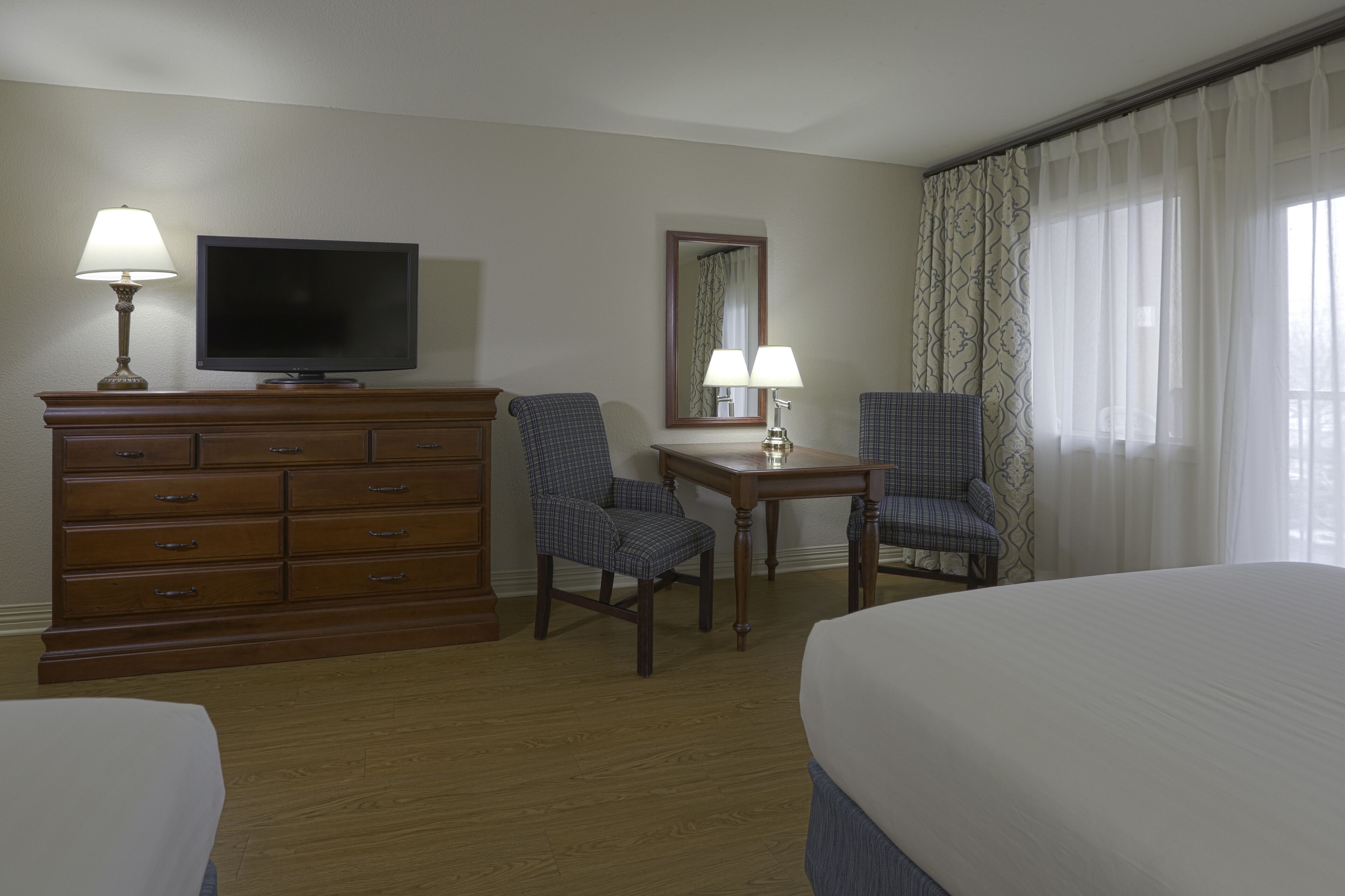 A hotel room with a bed, a TV, a desk with two chairs, a dresser, and a window with curtains.