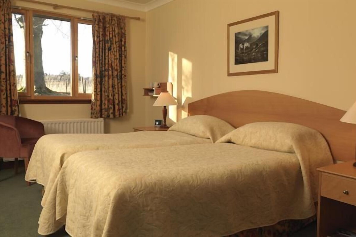A hotel room with two beds, a desk, a chair, a window with curtains, a lamp, and a framed picture on the wall.