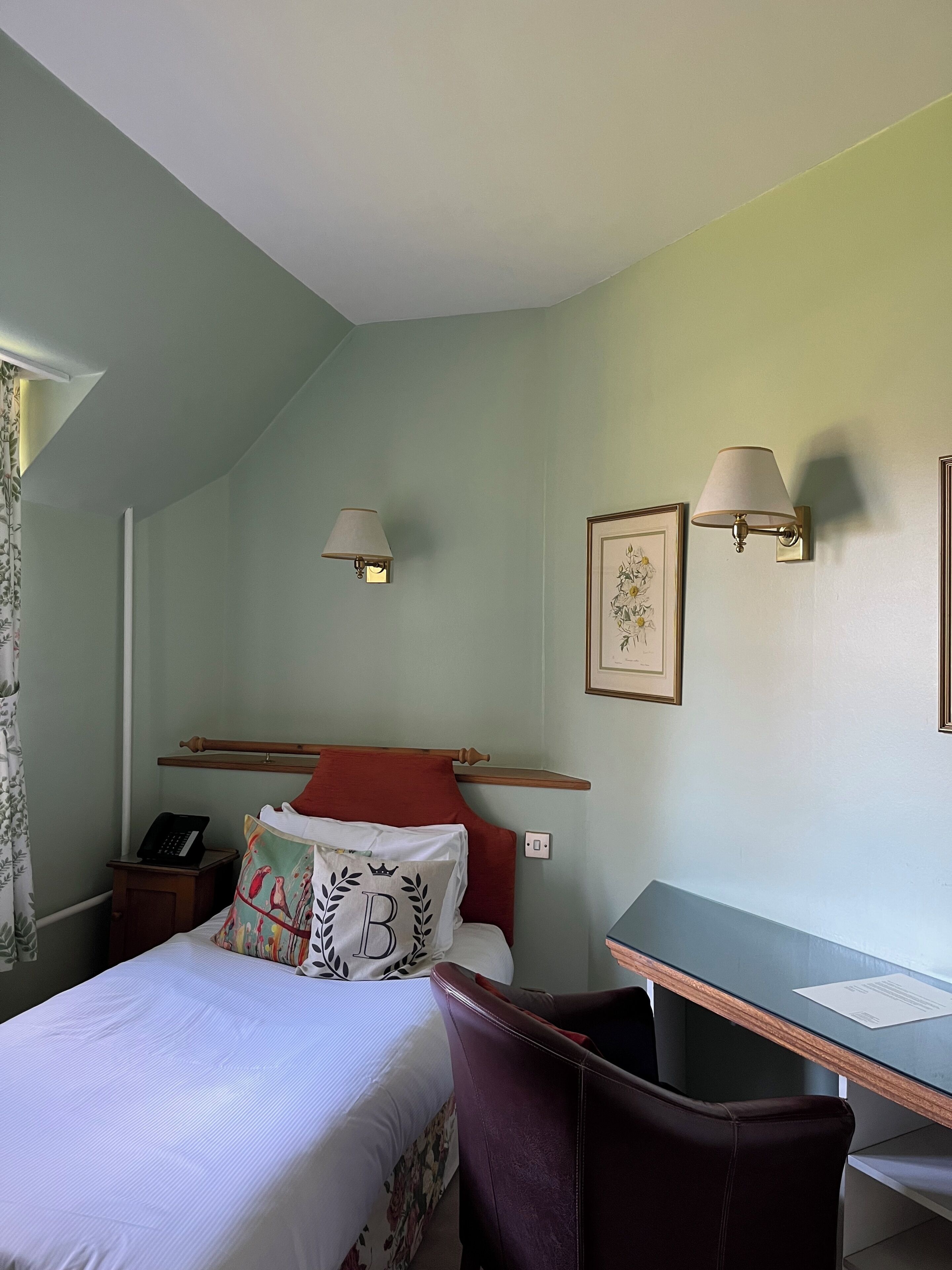 Ett hotellrum med en säng, ett skrivbord, två vägglampor, en telefon och en inramad tavla på väggen.