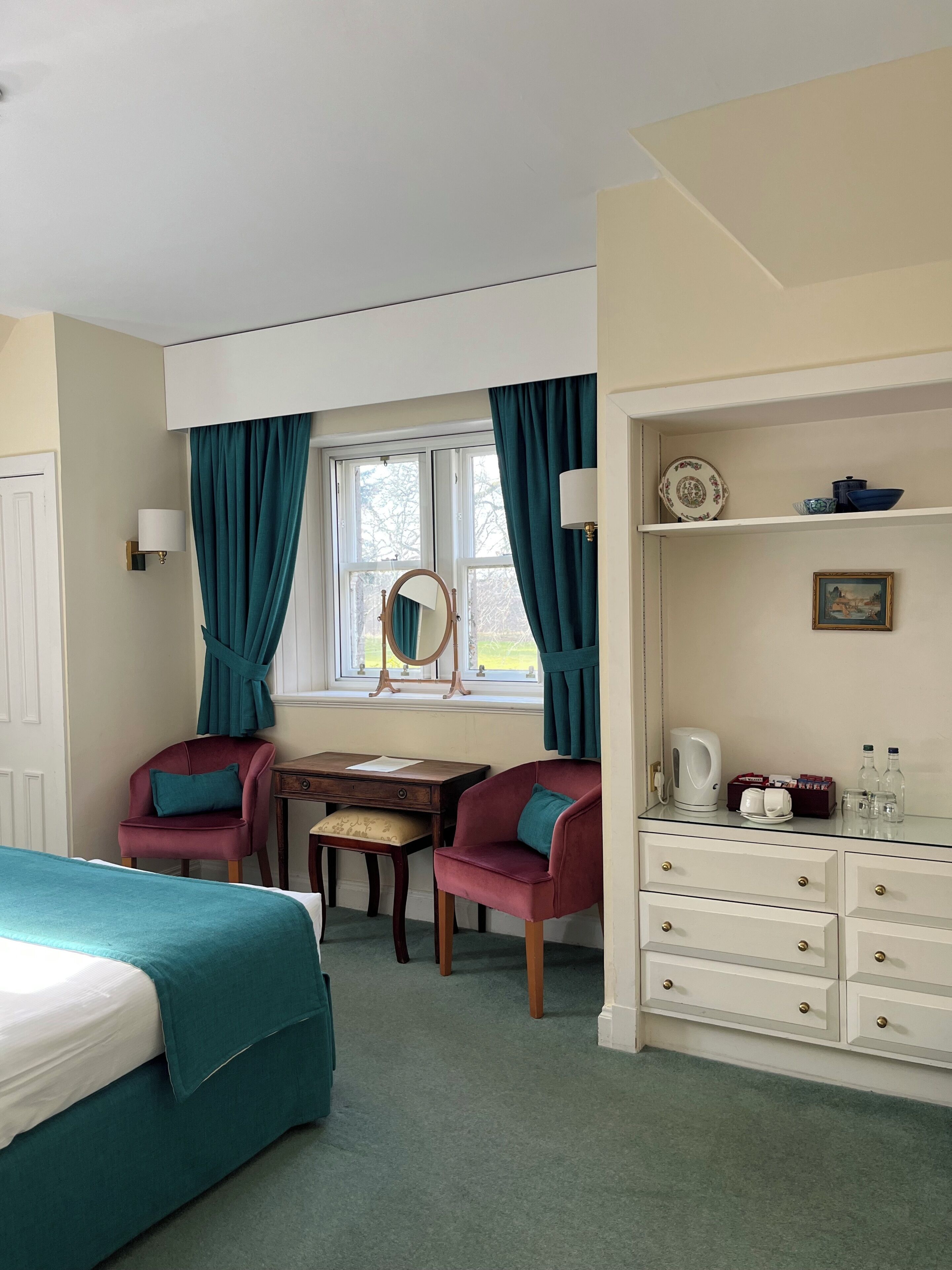 A hotel room with a bed, two chairs, a desk, a mirror, a dresser, and a window with curtains.