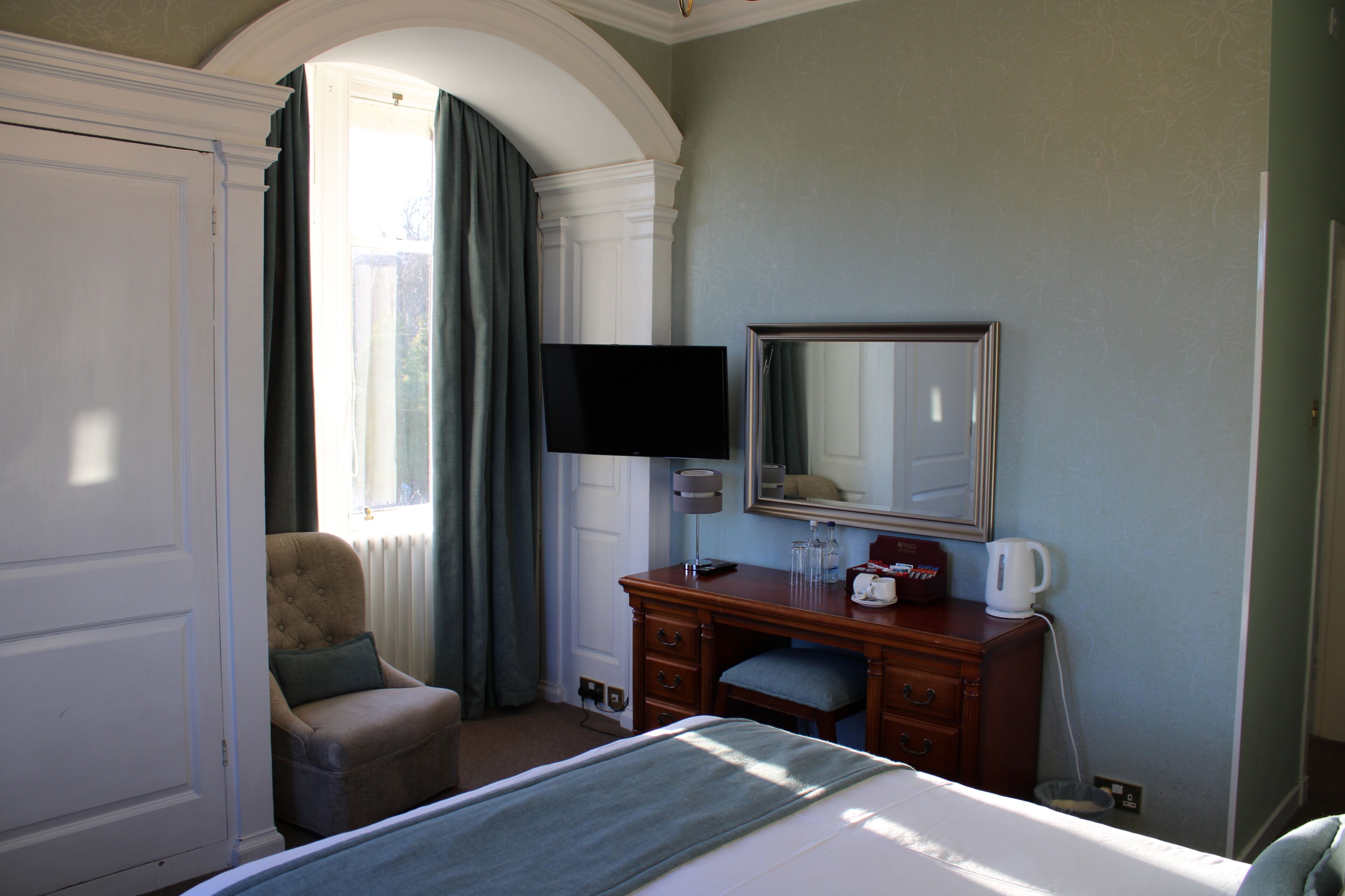 A hotel room with a bed, a desk with a flat-screen TV, a chair, a dresser, and a large mirror.