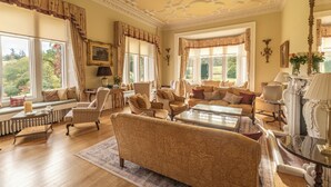 A spacious living room with a large sofa, multiple armchairs, a coffee table, and a fireplace. There are large windows with curtains, a radiator, and a chandelier. - Ballathie House Hotel (Perth)