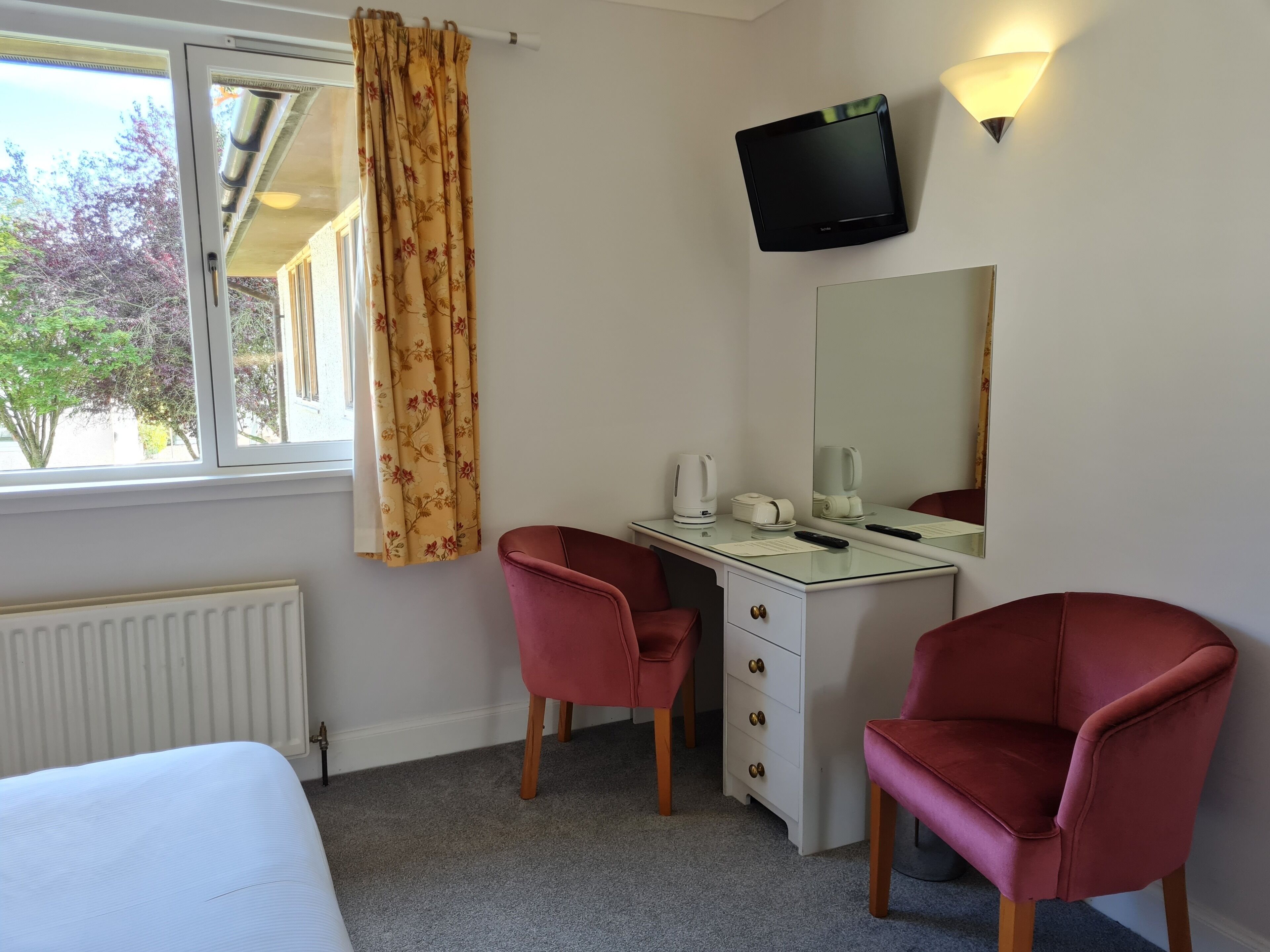 A hotel room with a bed, a desk with a chair, a television, a mirror, and a window with curtains.