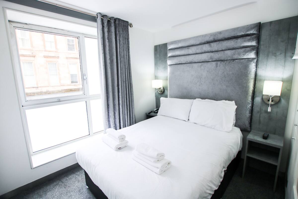 A hotel room with a large bed, two bedside tables with lamps, a window with curtains, and a view of buildings outside.