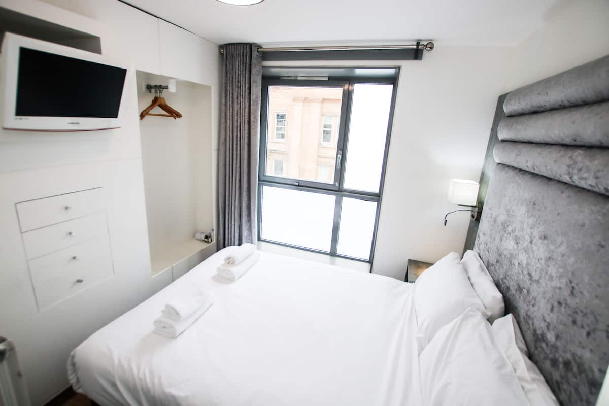 A hotel room with a bed, a TV mounted on the wall, a window with curtains, and a grey headboard.