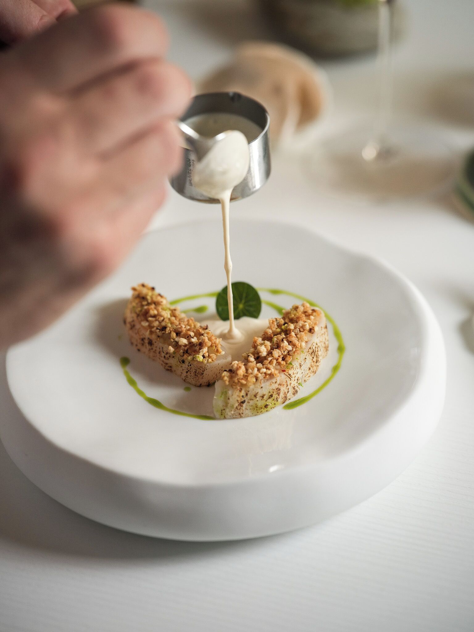A white plate with a food item topped with a crumbled topping and a dollop of sauce being poured on it.