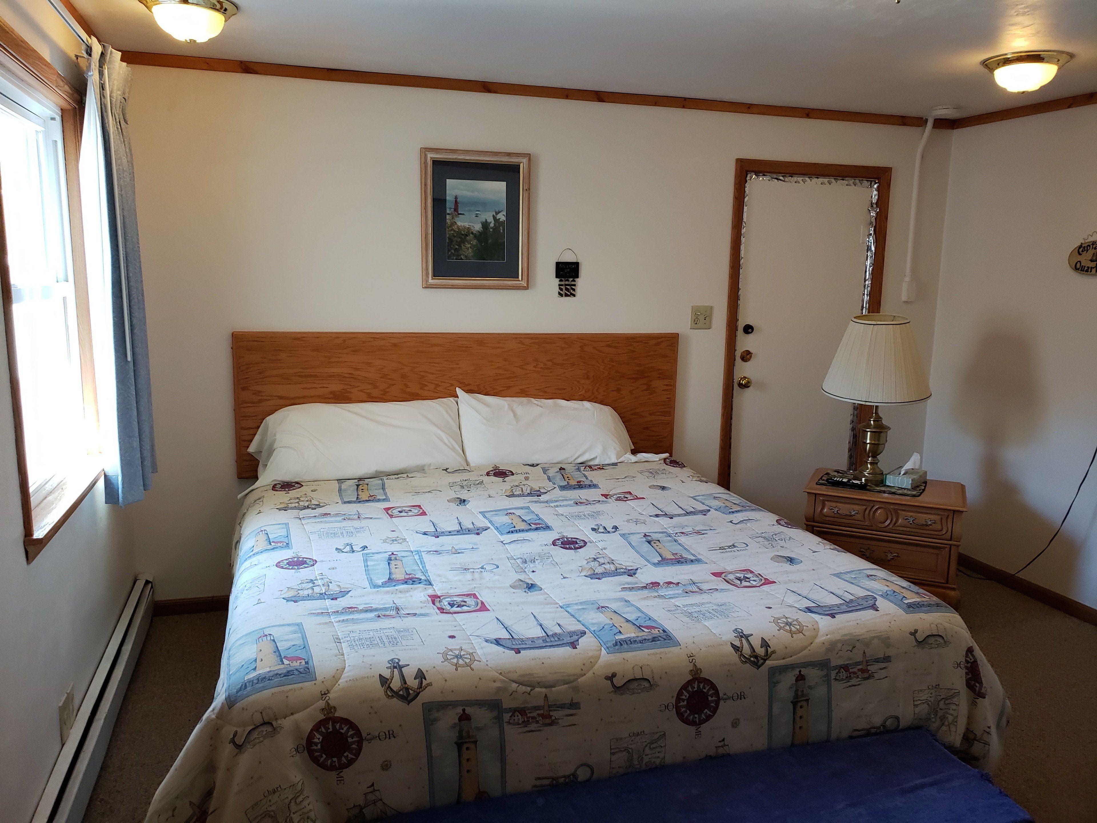 Un dormitorio con cabecera de madera, cama con colcha de estilo náutico, lámpara de noche y un cuadro enmarcado en la pared.