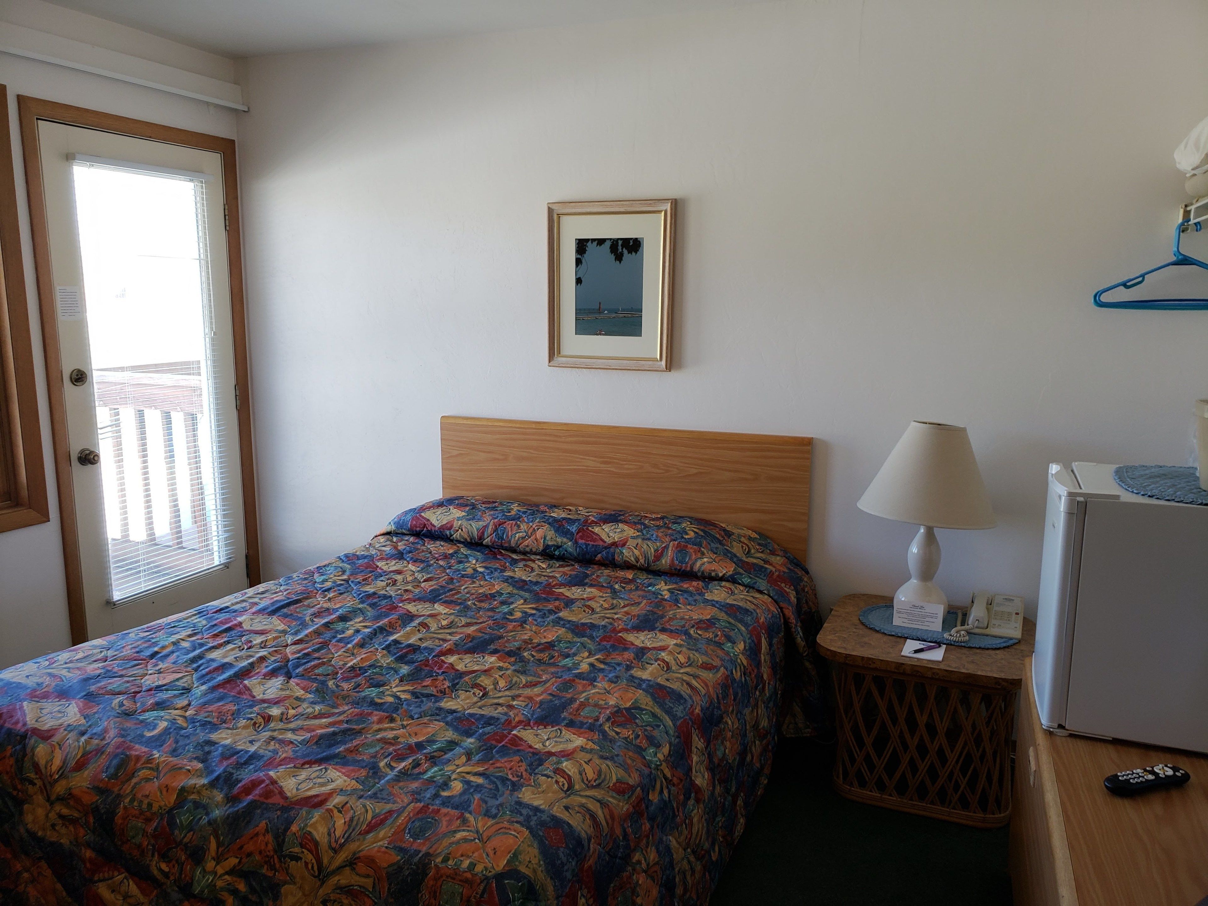 Een slaapkamer met een bed, een nachtkastje met een lamp, een koelkast en een deur met zonwering.